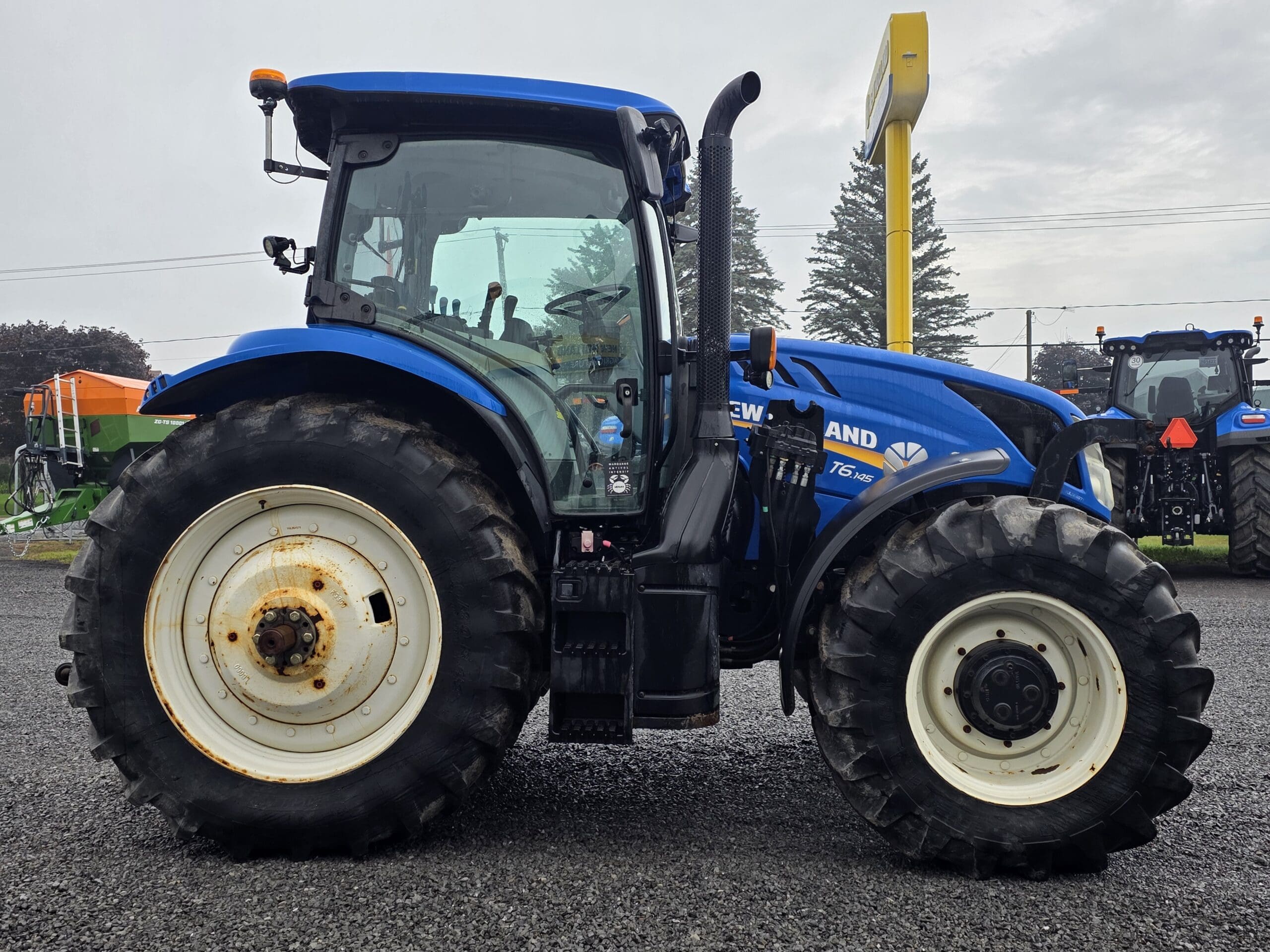 Un tracteur New Holland bleu avec de gros pneus à l'arrière et de petits pneus à l'avant est garé sur du gravier, avec du matériel agricole et des arbres visibles en arrière-plan sous un ciel nuageux.