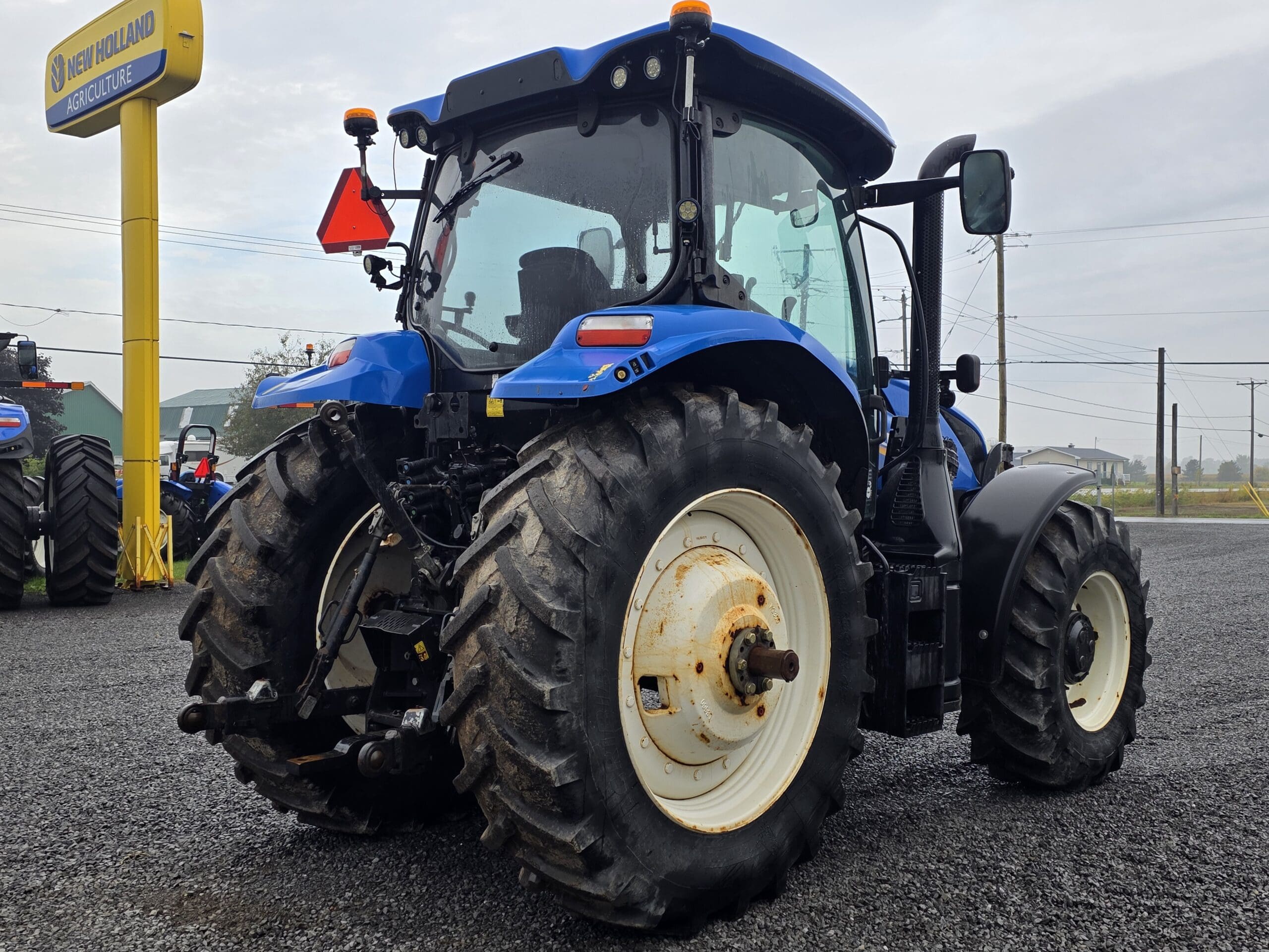 Un tracteur bleu New Holland est garé sur du gravier près d'un panneau jaune New Holland Agriculture, avec de gros pneus arrière et des pièces mécaniques visibles à l'arrière. Le ciel est couvert et il y a des lignes électriques à l'arrière-plan.