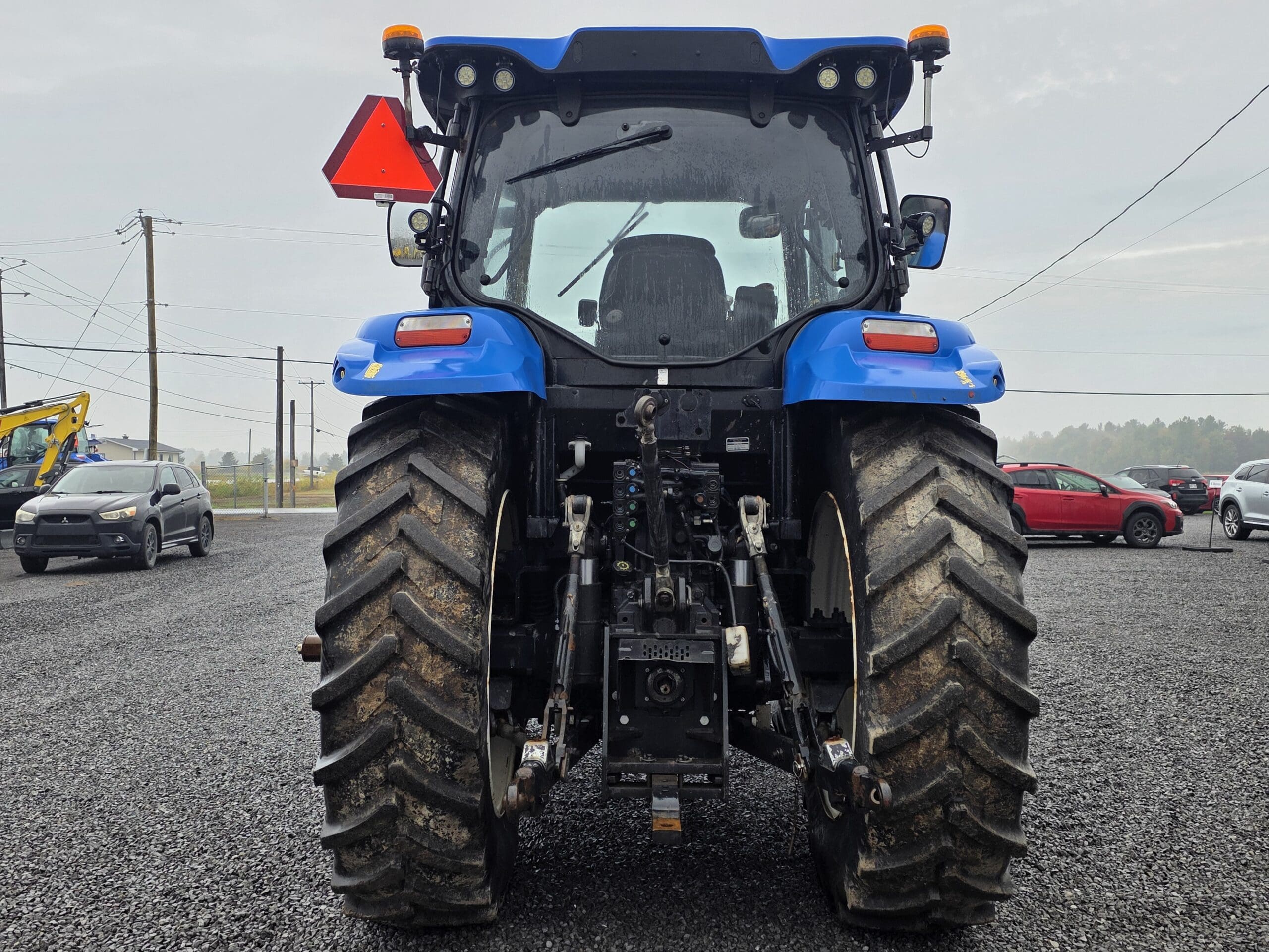 Vue arrière d'un tracteur bleu à gros pneus boueux garé sur du gravier. Un panneau triangulaire rouge indiquant qu'il s'agit d'un véhicule lent est fixé à l'arrière. Des voitures et des poteaux électriques sont visibles à l'arrière-plan.