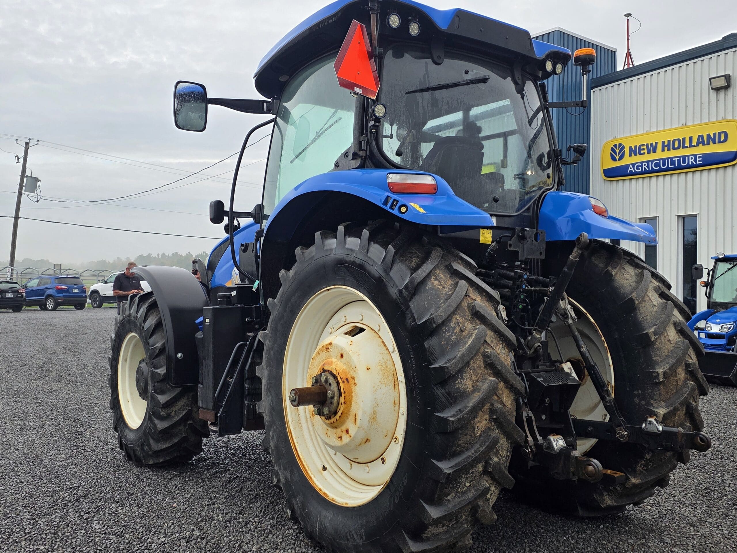 Un tracteur New Holland bleu avec de gros pneus arrière est garé à l'extérieur d'un concessionnaire New Holland Agriculture sur un terrain en gravier. Le tracteur présente un peu de rouille sur le moyeu de la roue. D'autres véhicules et bâtiments sont visibles à l'arrière-plan.