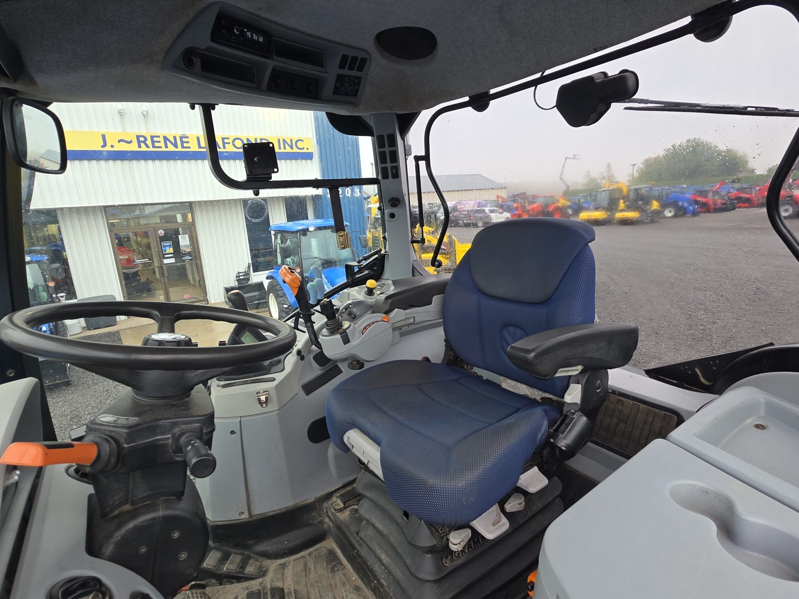 Intérieur d'une cabine de tracteur montrant un siège de conducteur bleu, un volant et diverses commandes, avec une vue d'un terrain de concession et de tracteurs visibles à travers les fenêtres avant et latérales.