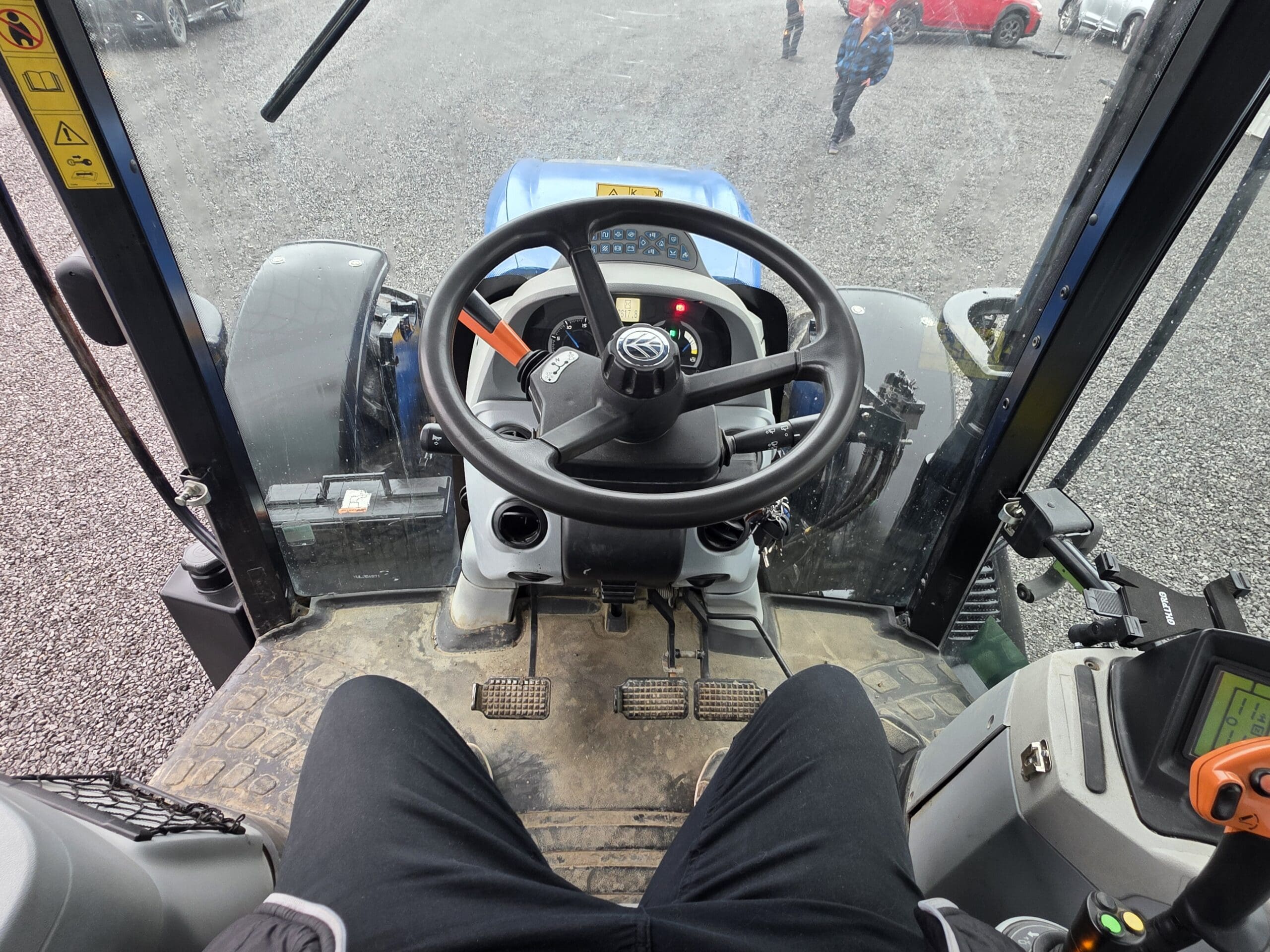 Vue de l'intérieur d'une cabine de tracteur montrant un volant, des pédales, des commandes sur le tableau de bord et les jambes d'une personne vêtue d'un pantalon noir. Le sol et quelques véhicules sont visibles à travers la vitre avant.