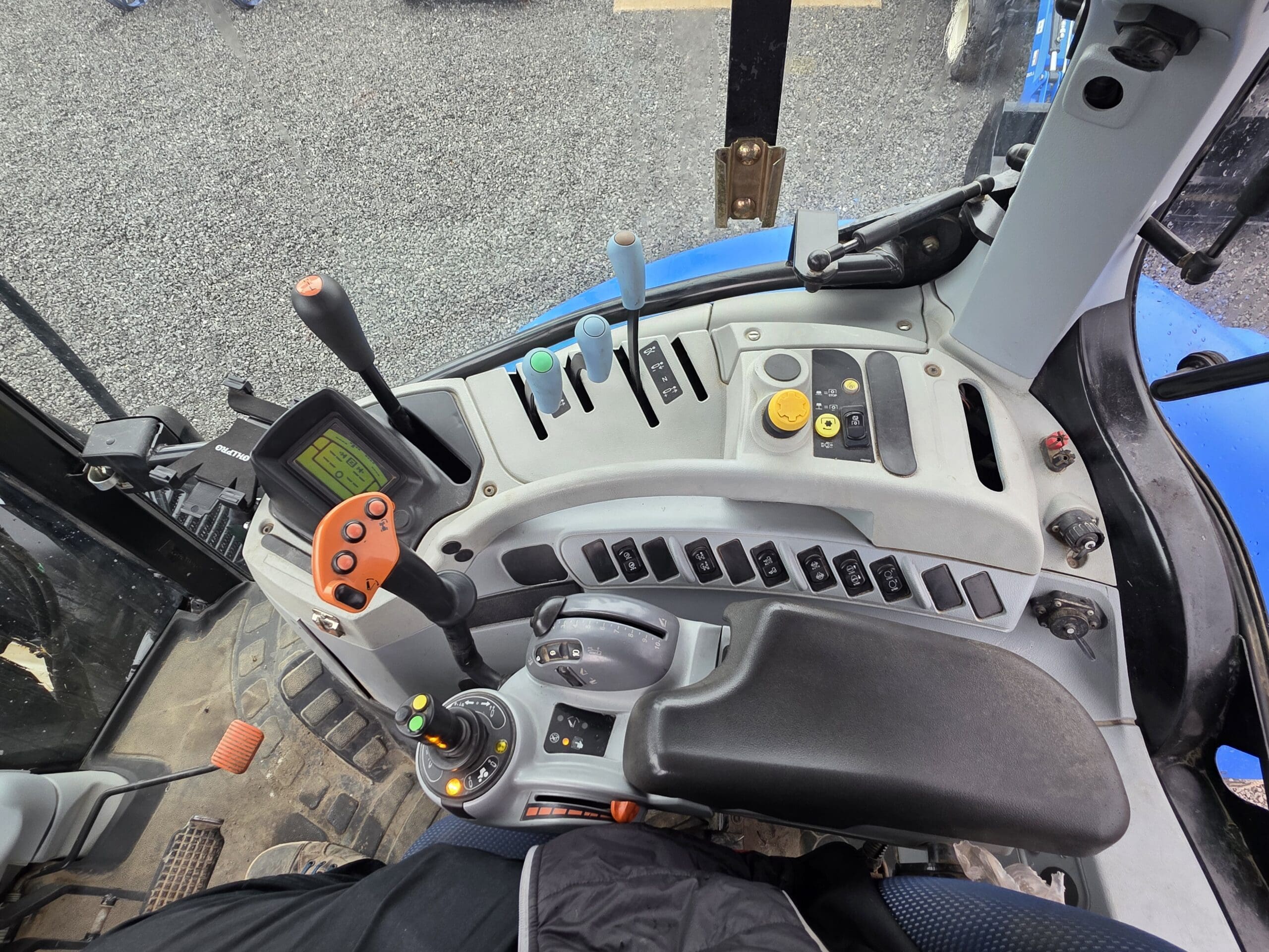 Vue de l'intérieur d'une cabine de tracteur montrant divers leviers de commande, boutons, interrupteurs, un écran d'affichage et un volant. La jambe de l'opérateur est partiellement visible en bas de l'image.