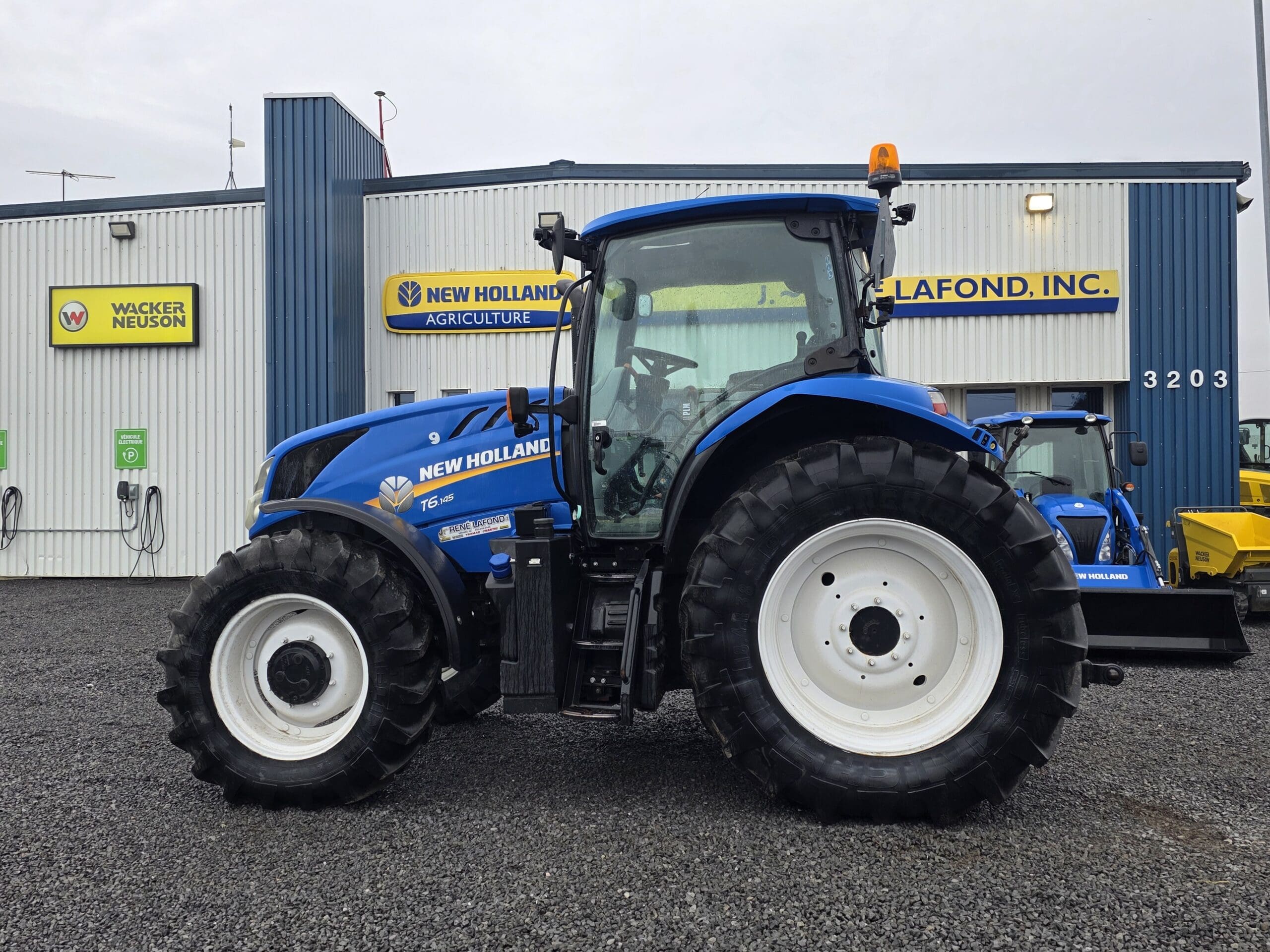 Un tracteur New Holland bleu est garé sur du gravier devant un bâtiment de Lafond, Inc. Des enseignes de l'entreprise sont visibles sur les murs extérieurs bleus et blancs du bâtiment.