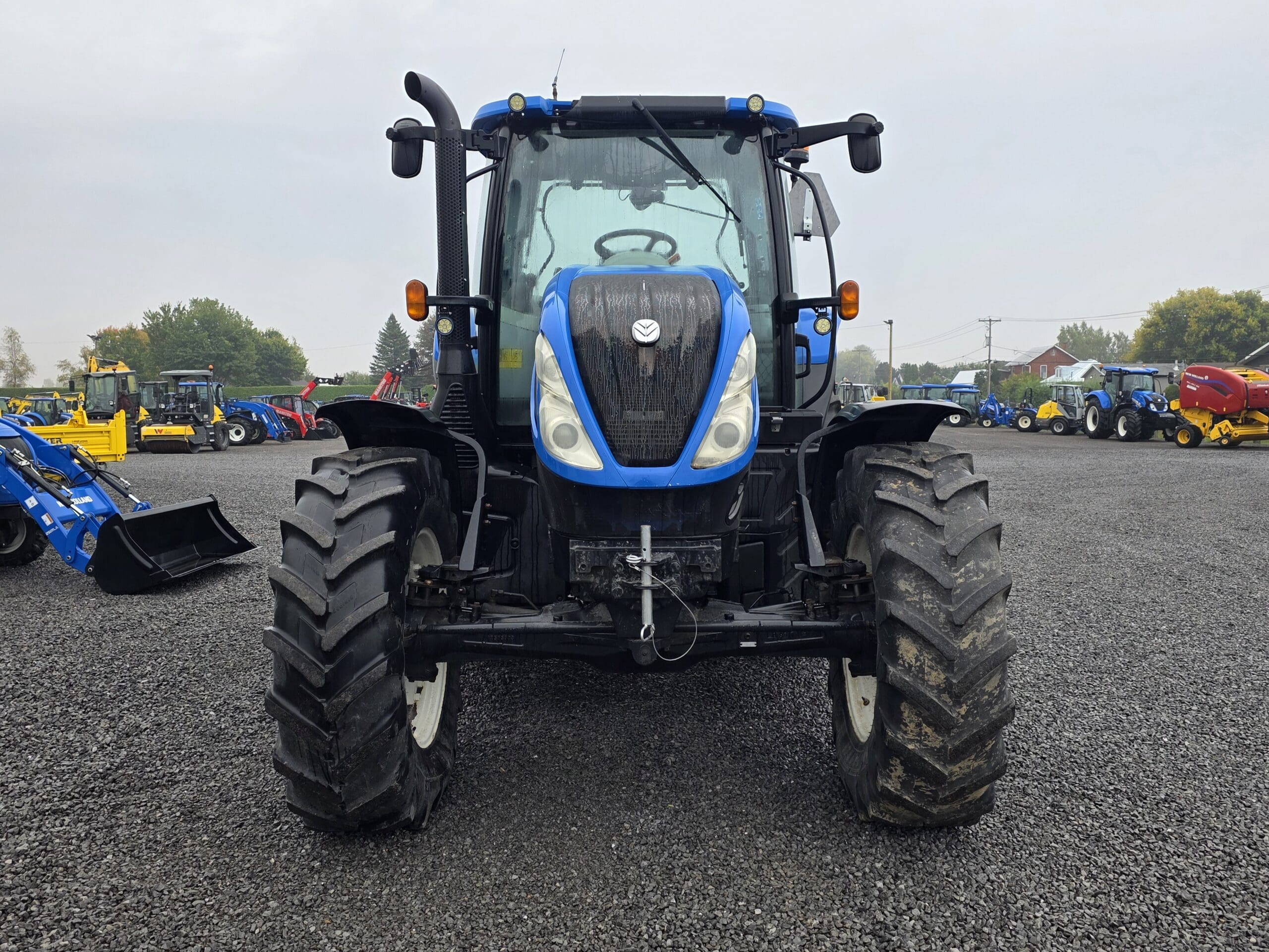 Un tracteur bleu est garé sur un terrain en gravier, face à la caméra. D'autres équipements et machines agricoles sont visibles à l'arrière-plan sous un ciel couvert.