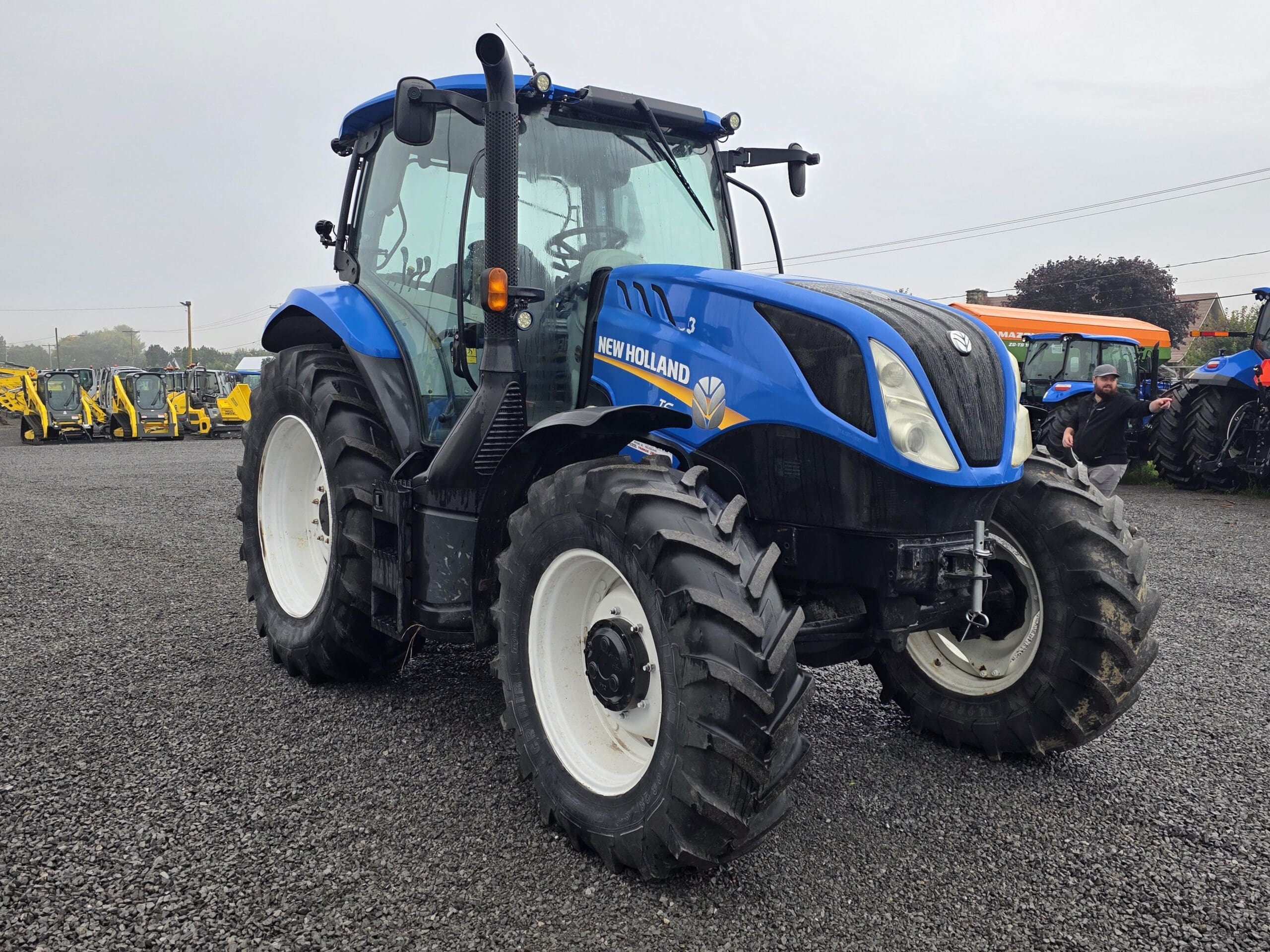 Un tracteur New Holland bleu est garé sur un terrain en gravier, avec d'autres tracteurs et machines visibles à l'arrière-plan. Le ciel est couvert et une personne est aperçue près d'un autre tracteur à droite.