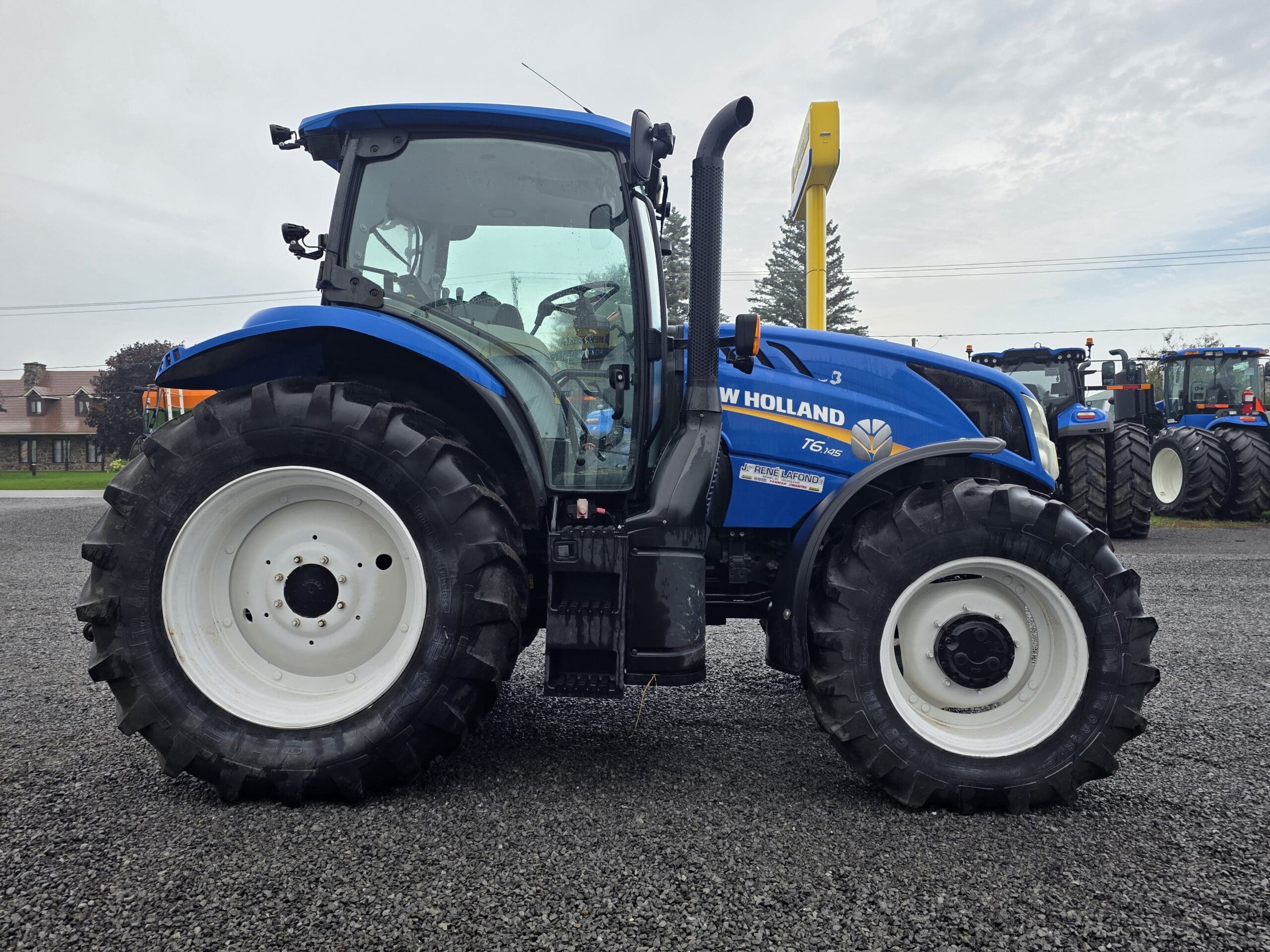 Un tracteur New Holland T5 bleu est garé sur une surface de gravier, avec de gros pneus noirs et un panneau de signalisation jaune en arrière-plan, sous un ciel nuageux.