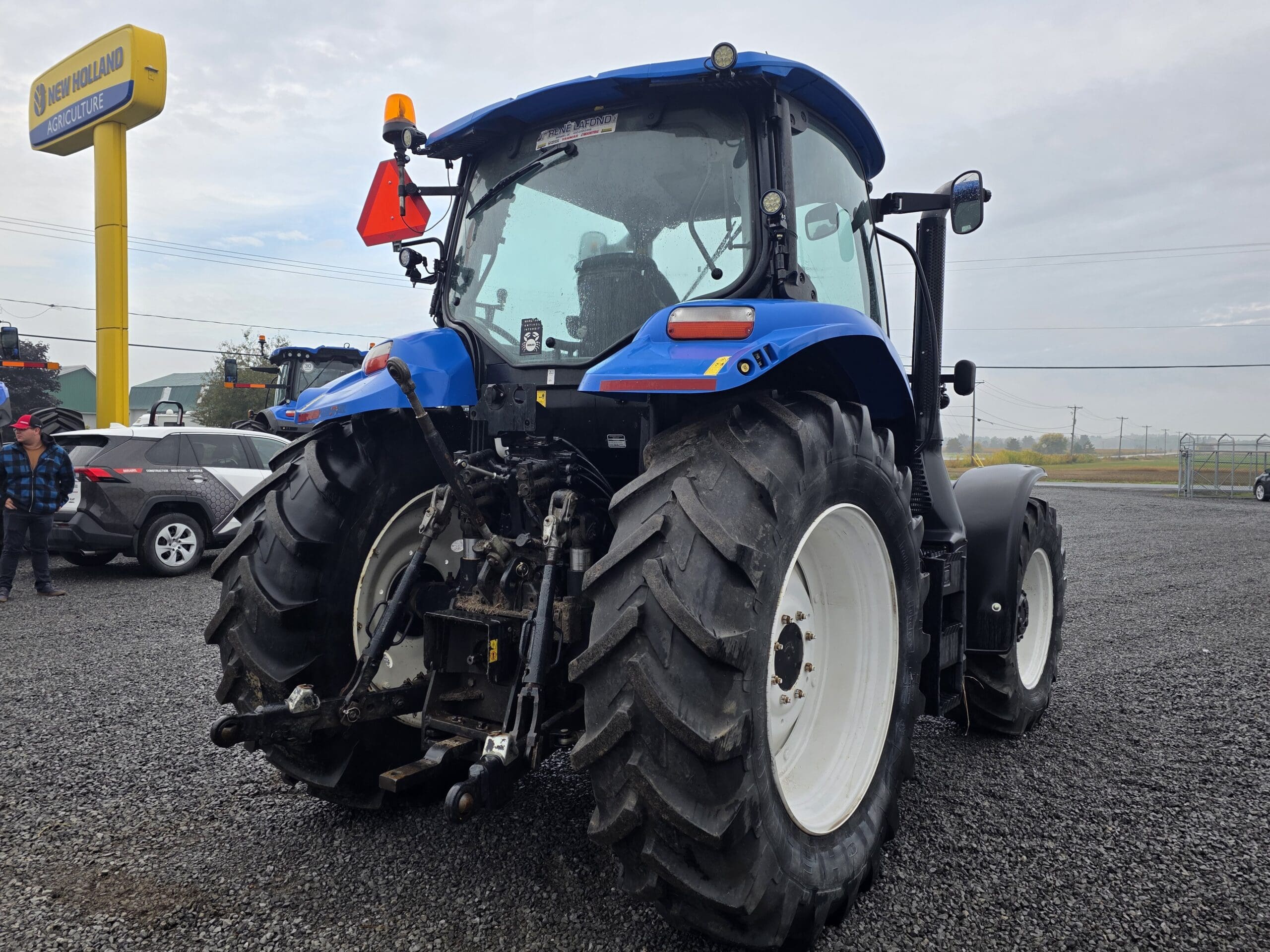 Un tracteur New Holland bleu est garé sur du gravier près d'une enseigne de concessionnaire, avec de gros pneus arrière et du matériel agricole attaché à l'arrière. D'autres véhicules et personnes sont visibles à l'arrière-plan.