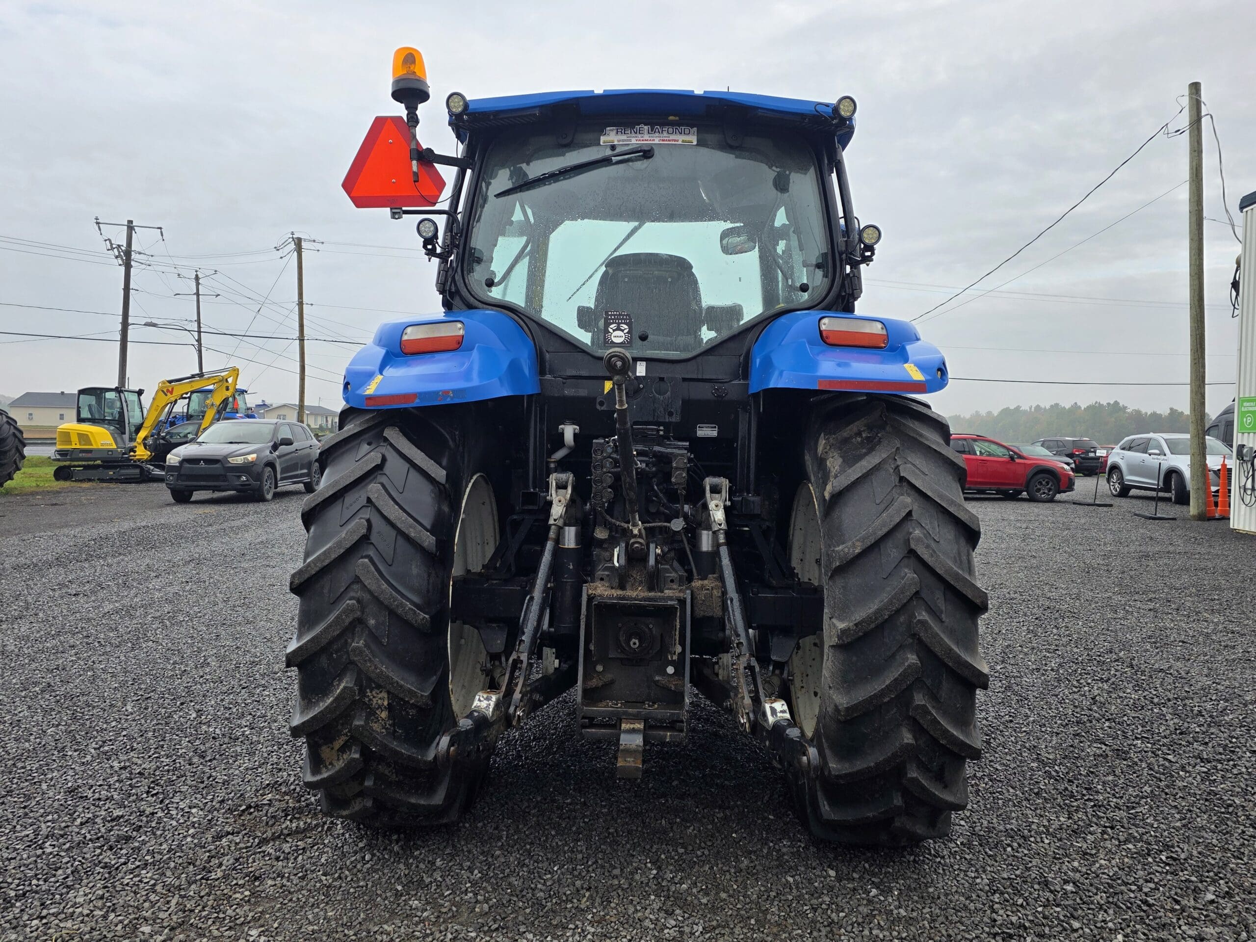 Un tracteur bleu vu de l'arrière, garé sur un terrain en gravier avec d'autres véhicules et équipements de construction en arrière-plan. Le tracteur est équipé de gros pneus, d'un feu d'avertissement et d'un panneau triangulaire réfléchissant.
