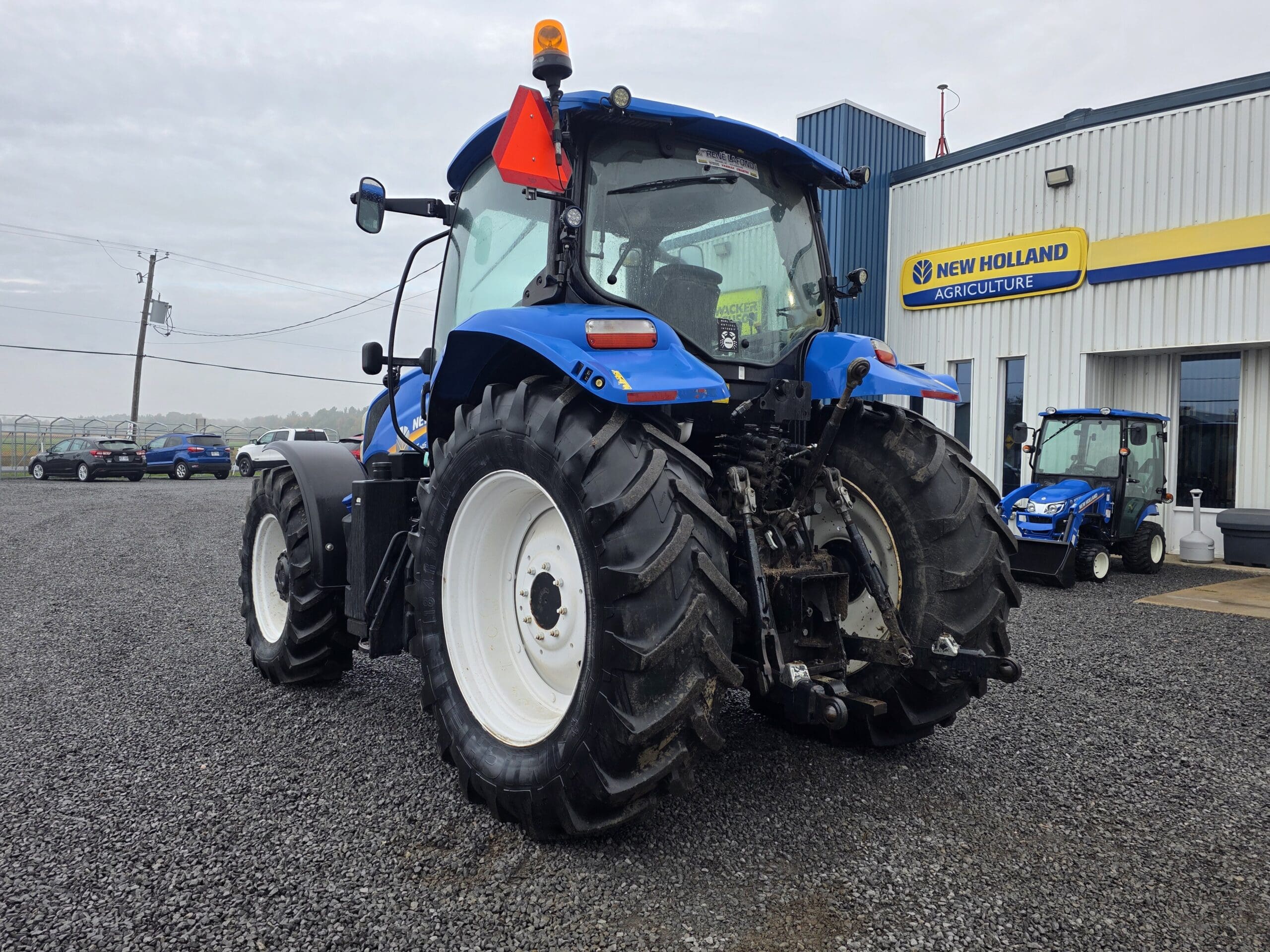 Un tracteur New Holland bleu est garé sur du gravier à l'extérieur d'une concession New Holland Agriculture, un autre petit tracteur bleu étant visible près de l'entrée du bâtiment.