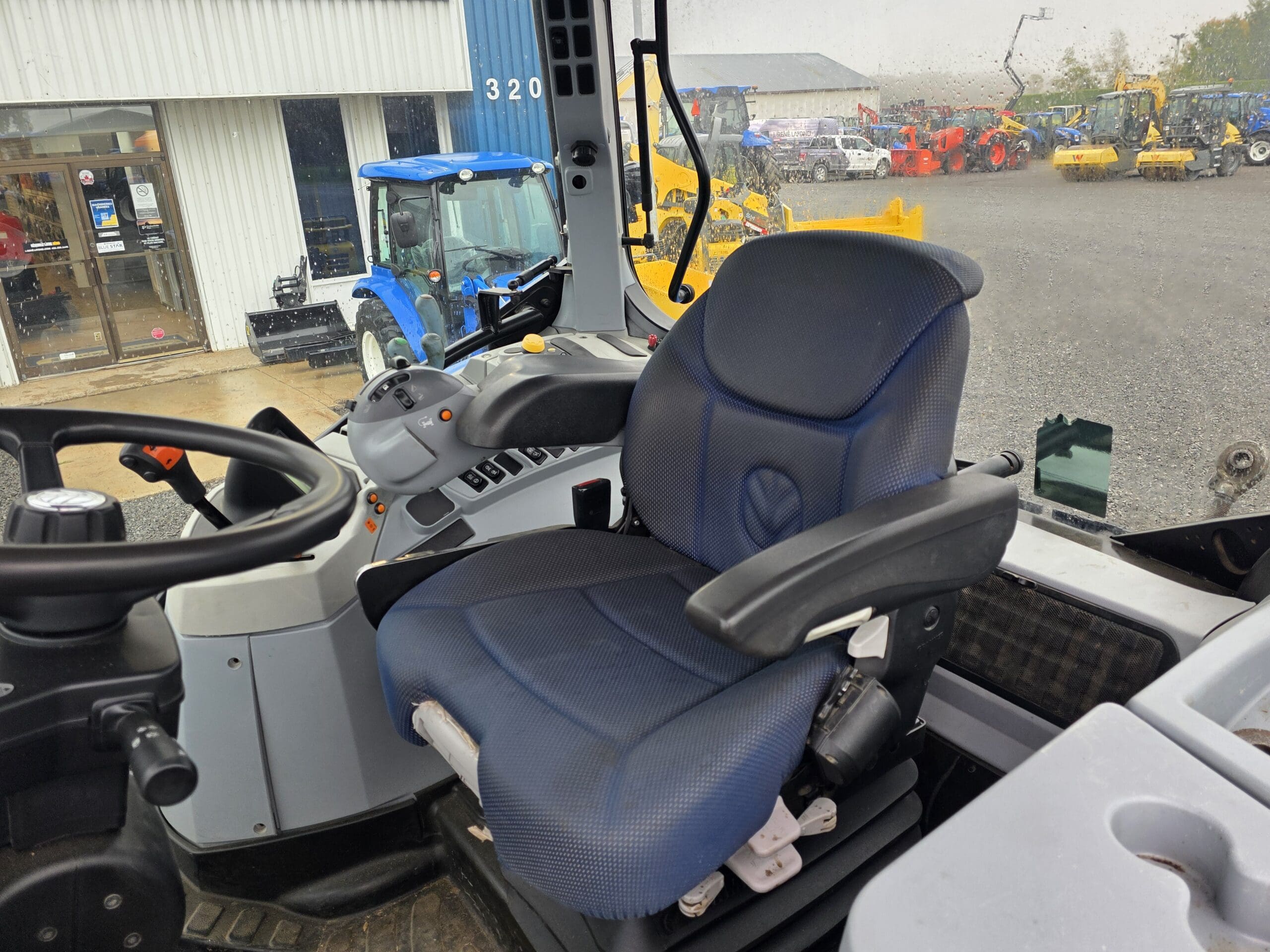 Vue intérieure d'une cabine de tracteur montrant un siège de conducteur à coussin bleu, un volant, un panneau de commande et une grande fenêtre avec une vue sur l'équipement et les véhicules à l'extérieur par une journée pluvieuse.