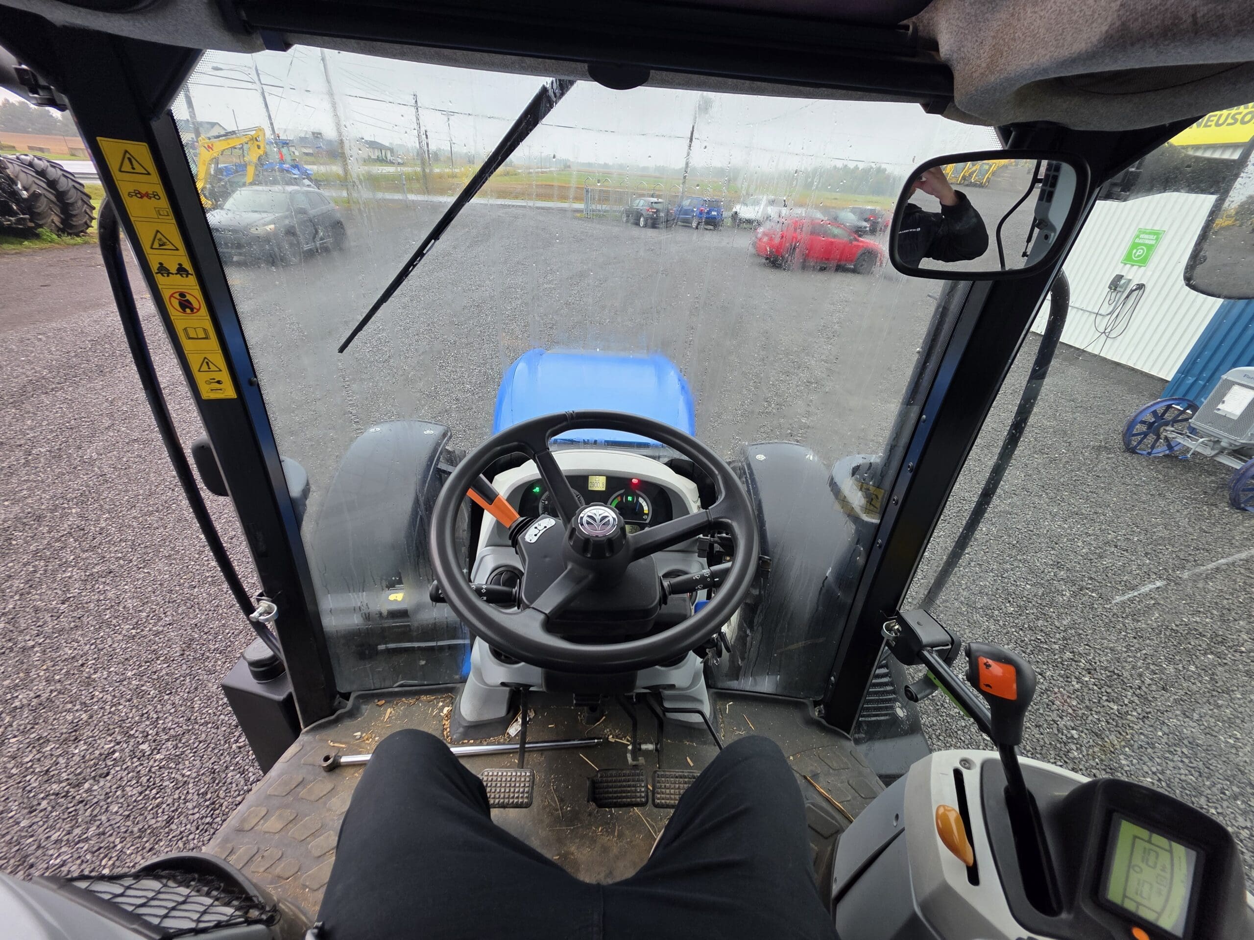 Vue de l'intérieur d'une cabine de tracteur, montrant le volant, les commandes du tableau de bord et les jambes du conducteur. Le pare-brise révèle un terrain en gravier, des véhicules en stationnement et des bâtiments industriels à l'extérieur.
