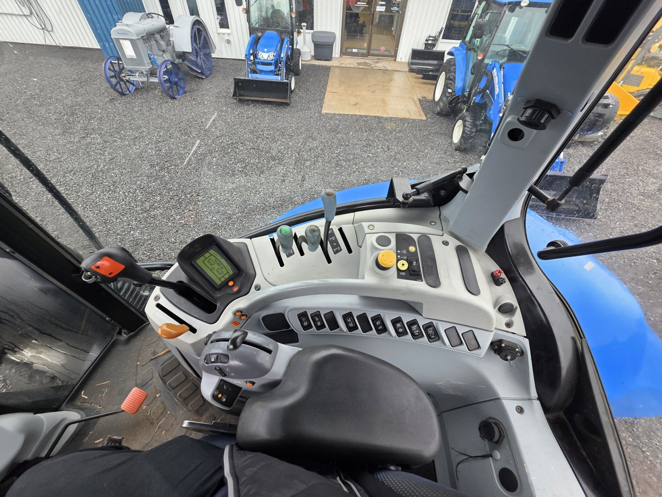 Vue de l'intérieur d'une cabine de tracteur moderne, montrant le siège du conducteur, le panneau de commande avec des boutons, des leviers et un écran numérique. Plusieurs tracteurs bleus et machines anciennes sont visibles à l'extérieur à travers le pare-brise.