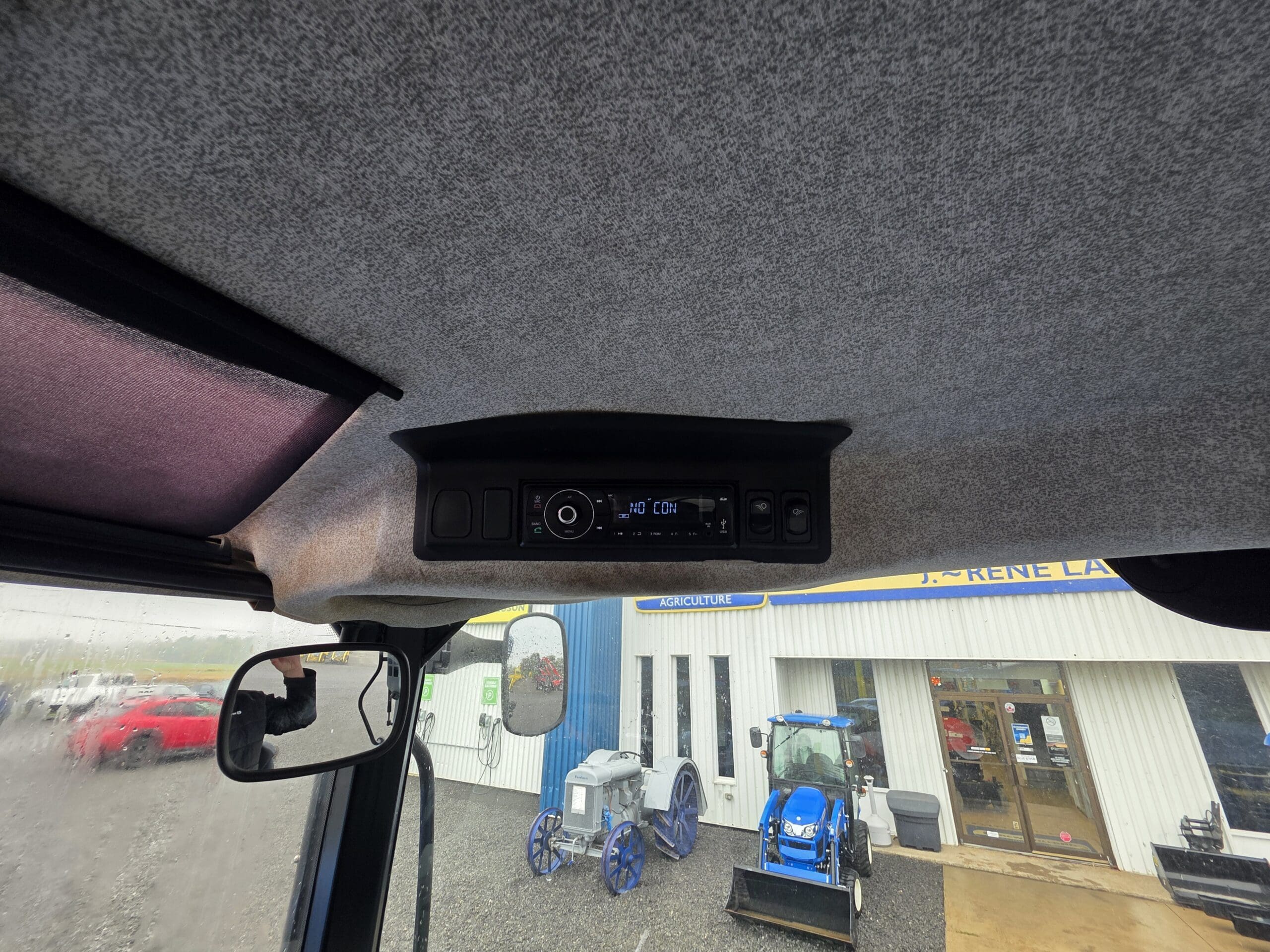 Vue de l'intérieur d'une cabine de véhicule montrant une console radio au plafond. À l'extérieur de la fenêtre, on aperçoit des tracteurs, un véhicule rouge et un bâtiment blanc avec une enseigne bleue et jaune. La pluie est visible sur la fenêtre.