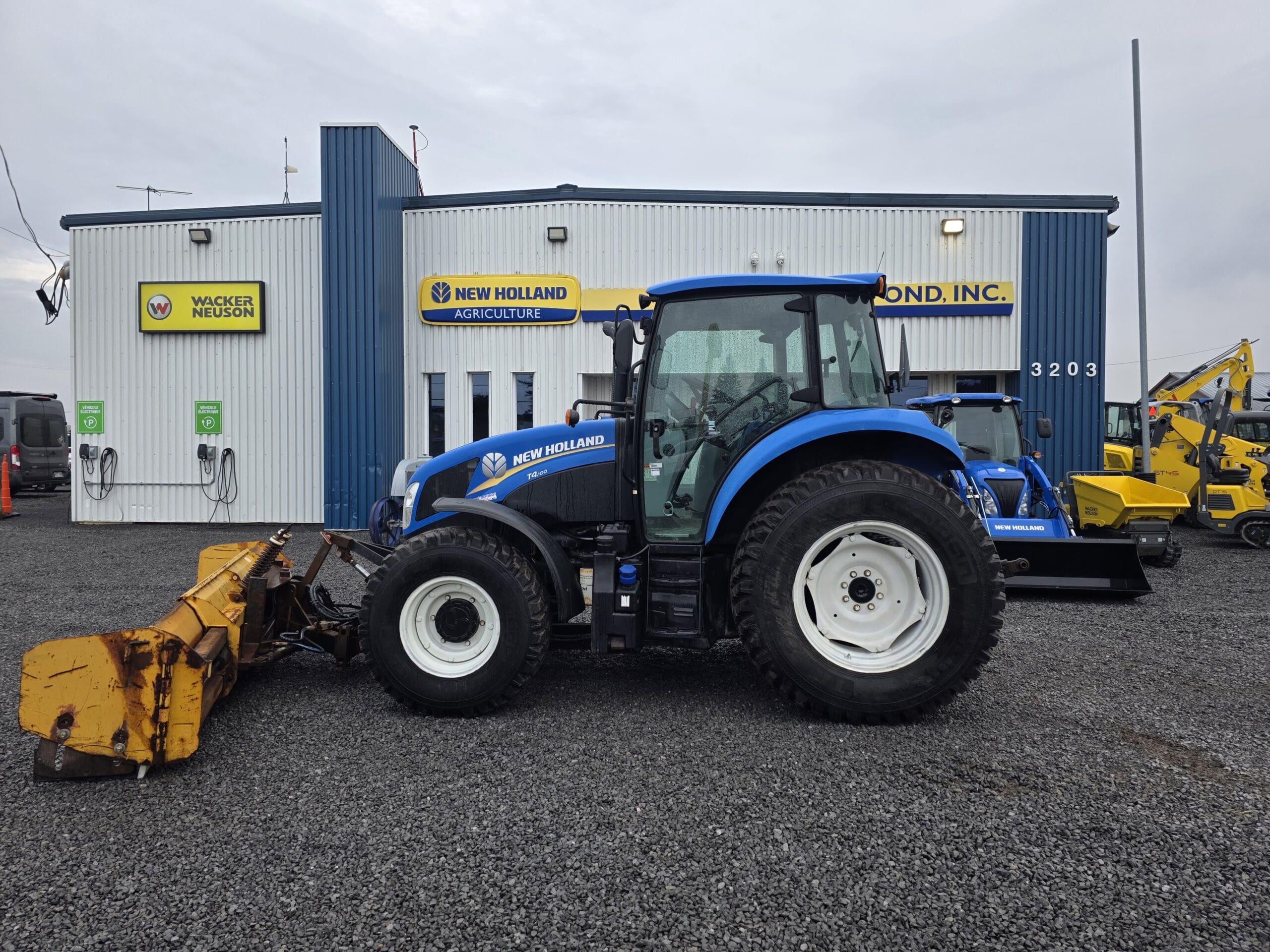 Un tracteur New Holland bleu avec un outil frontal jaune est garé sur du gravier devant le bâtiment d'un concessionnaire New Holland Agriculture. D'autres machines et enseignes sont visibles à l'arrière-plan.