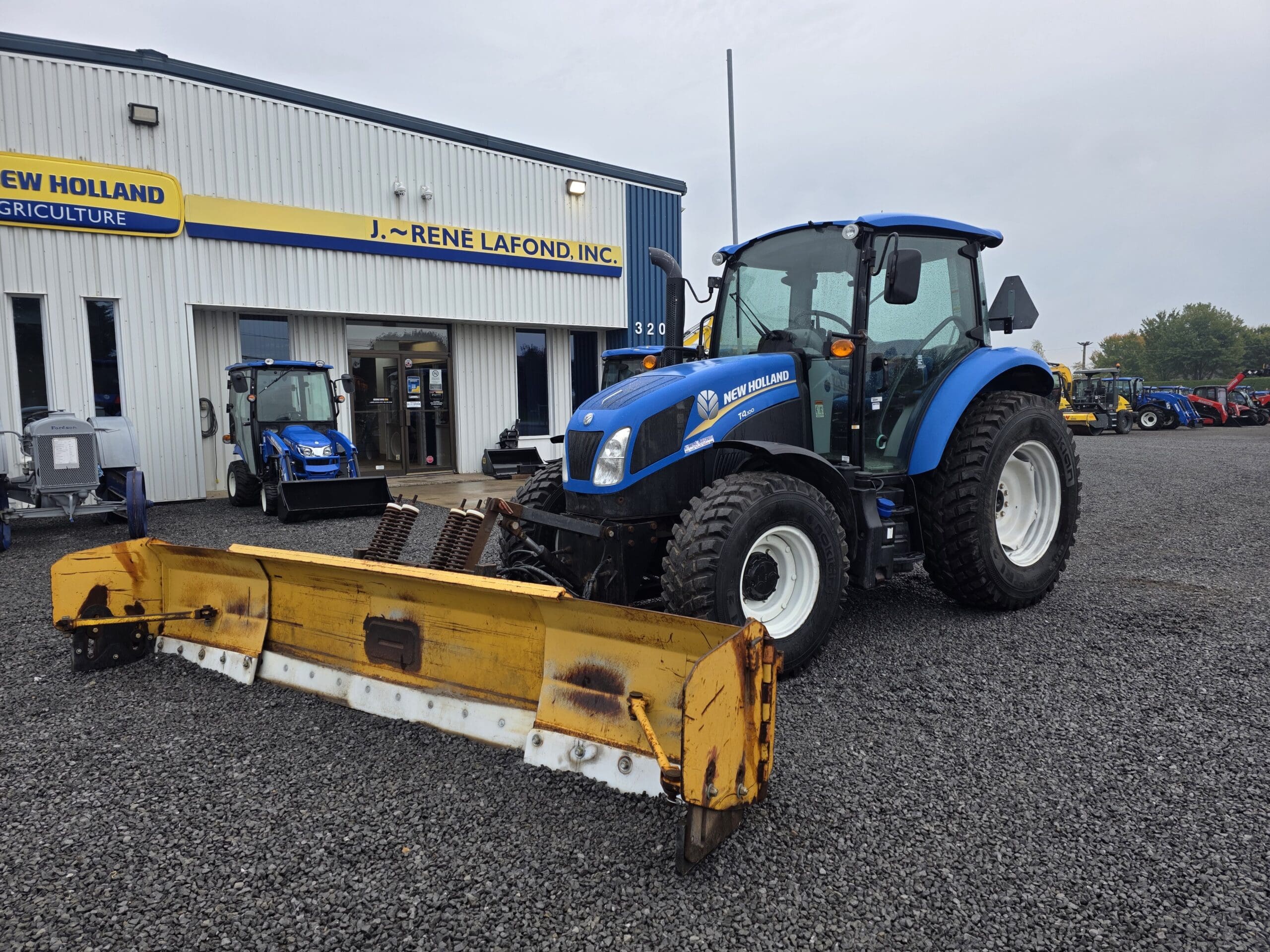 Un tracteur New Holland bleu avec un outil frontal jaune est garé sur du gravier à l'extérieur d'un bâtiment de la concession New Holland Agriculture. Un autre véhicule bleu est visible près de l'entrée.