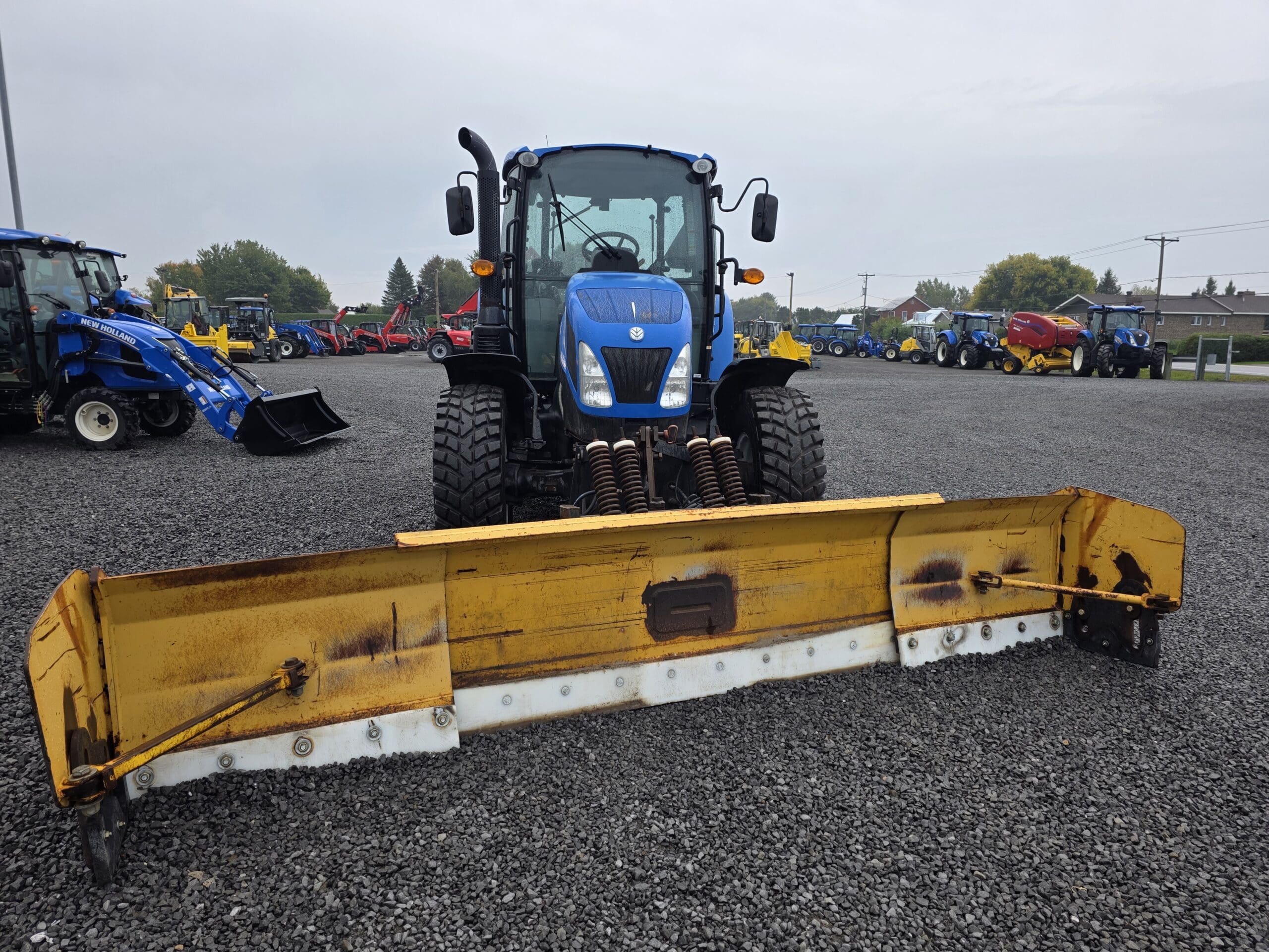 Un tracteur bleu à gros pneus est garé sur du gravier, équipé d'une large lame de déneigement jaune à l'avant. D'autres tracteurs et équipements sont visibles à l'arrière-plan par temps couvert.