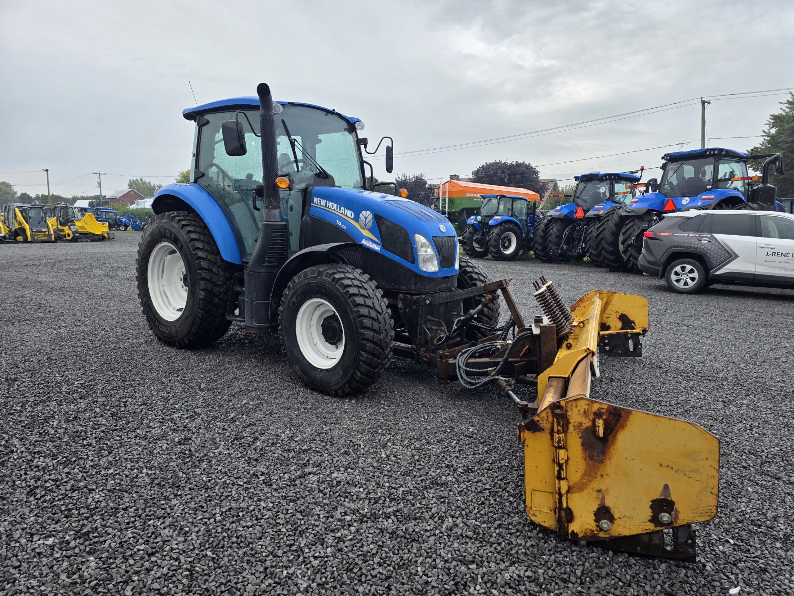 Un tracteur New Holland bleu avec un grand outil frontal jaune est garé sur un terrain en gravier. D'autres tracteurs et véhicules sont visibles à l'arrière-plan sous un ciel nuageux.