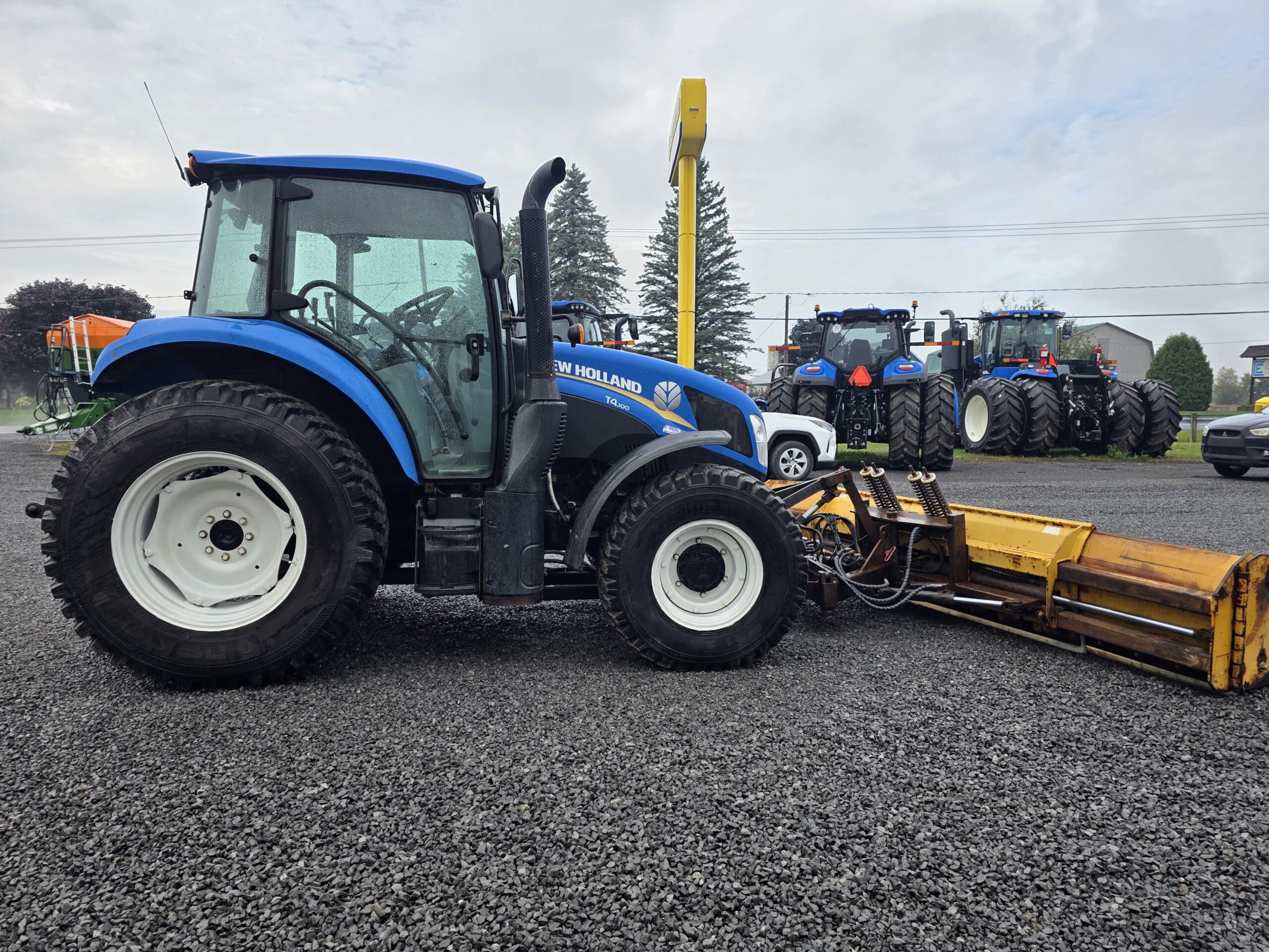 Un tracteur New Holland bleu avec de gros pneus est garé sur du gravier, attaché à un équipement jaune. D'autres tracteurs et une voiture sont visibles à l'arrière-plan, ainsi que des arbres et un ciel nuageux.