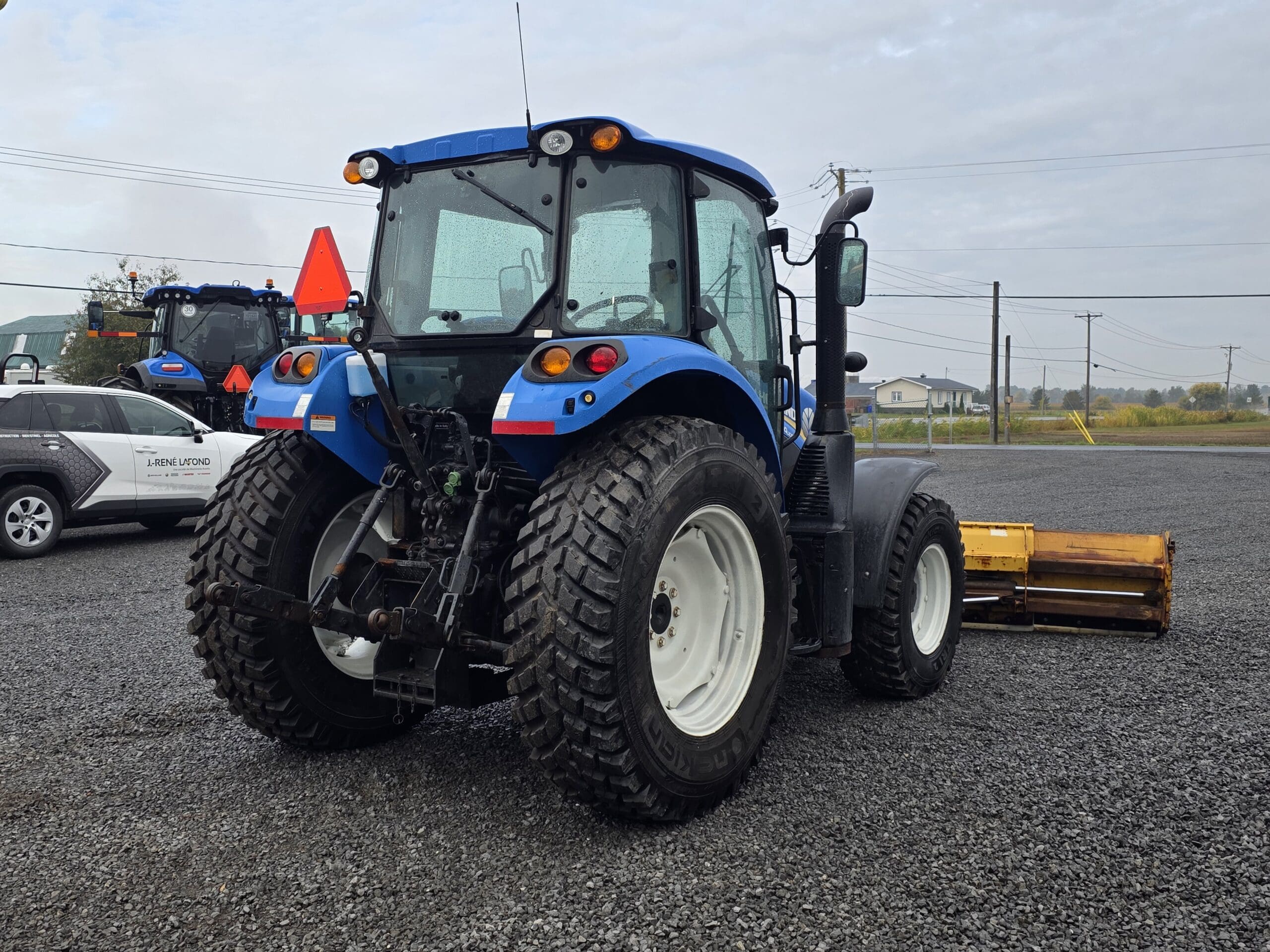 Un tracteur bleu à gros pneus est garé sur un terrain en gravier à côté d'un véhicule blanc. Des lignes électriques et des bâtiments sont visibles à l'arrière-plan sous un ciel nuageux.