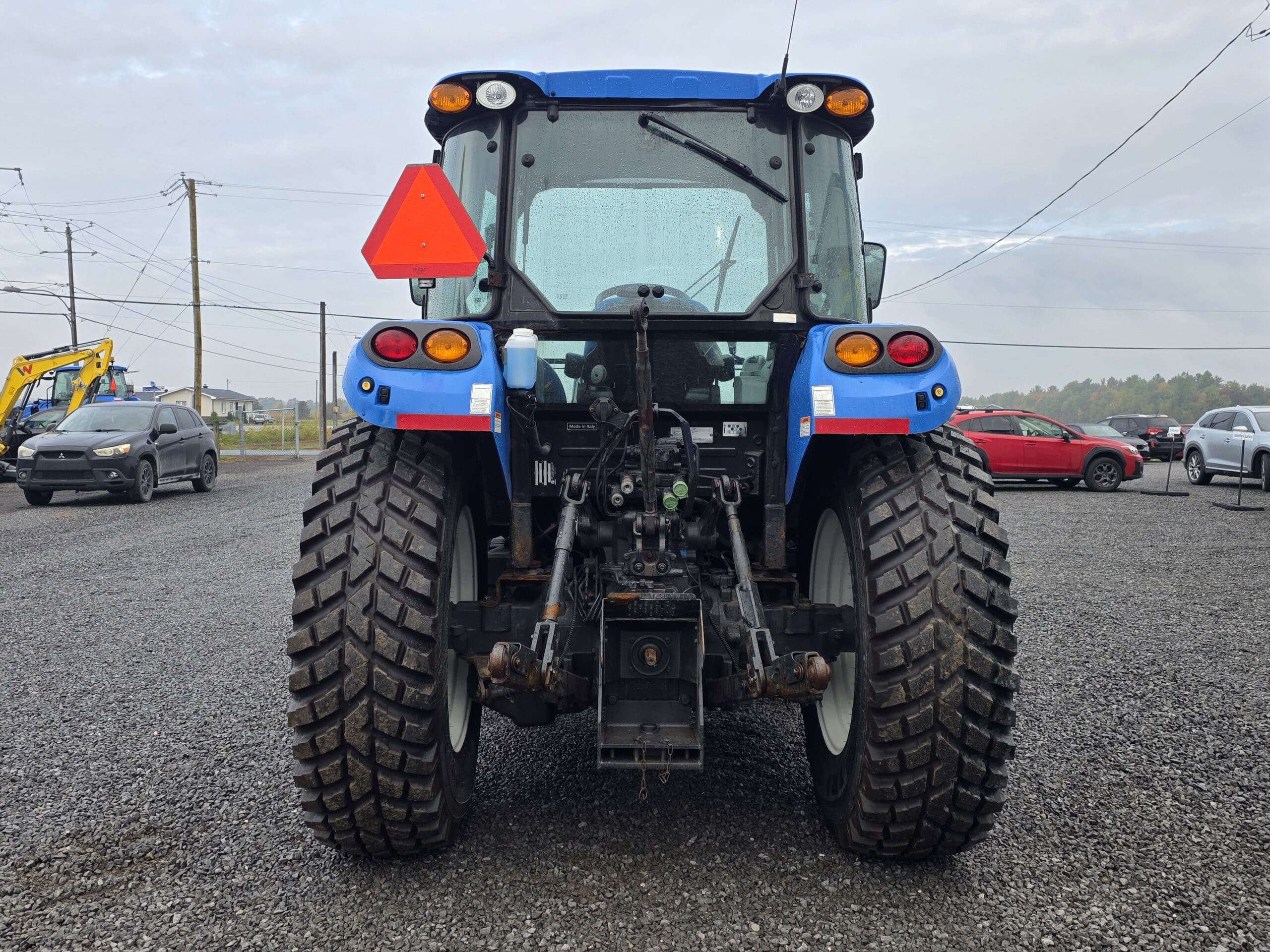 Un tracteur bleu vu de l'arrière, garé sur du gravier, avec de gros pneus, un panneau triangulaire rouge et divers véhicules visibles en arrière-plan sous un ciel nuageux.