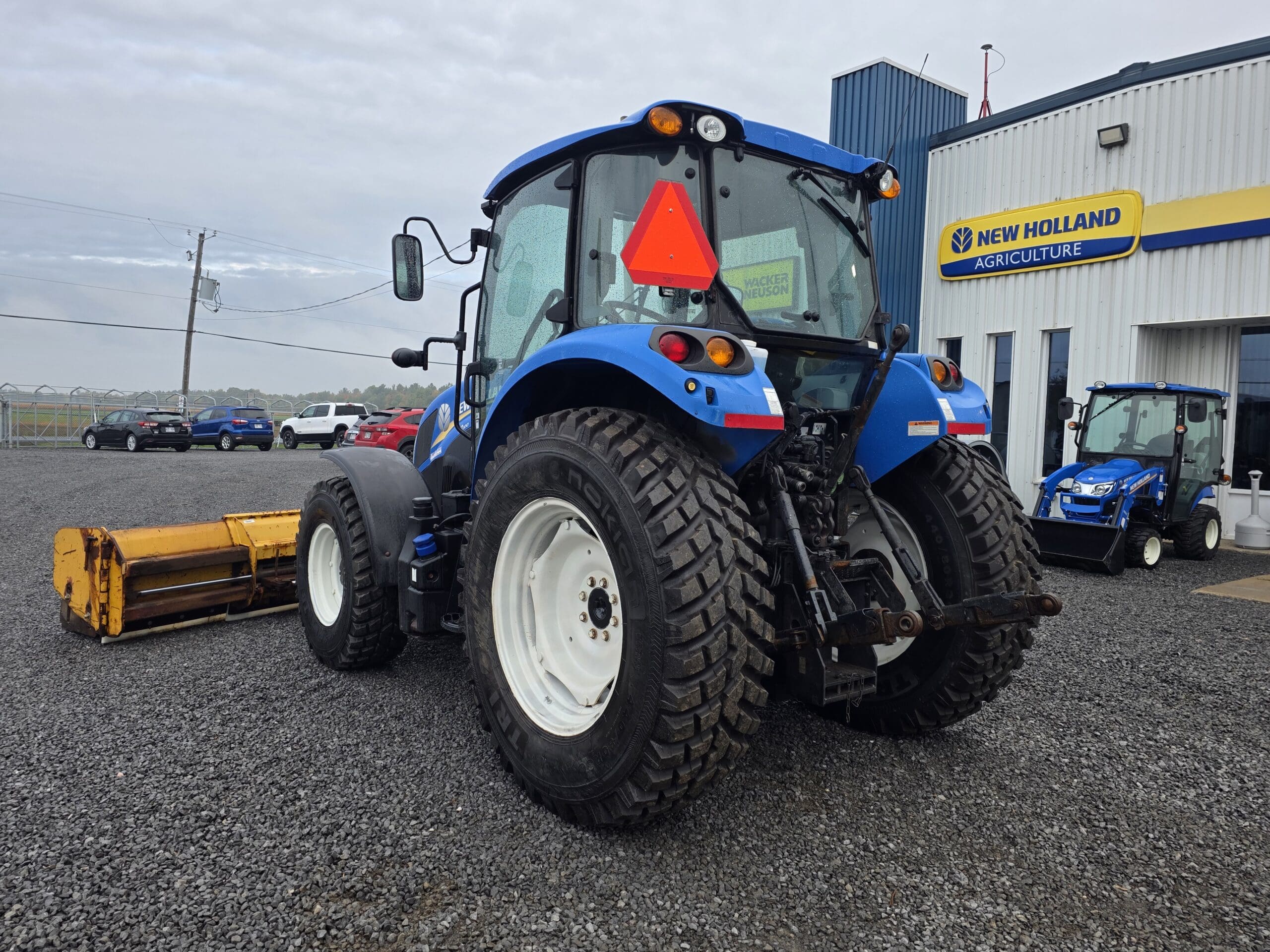 Un tracteur bleu New Holland avec de gros pneus est garé sur du gravier à l'extérieur d'un bâtiment New Holland Agriculture, avec un accessoire agricole jaune sur le sol derrière lui. Plusieurs véhicules se trouvent à l'arrière-plan.