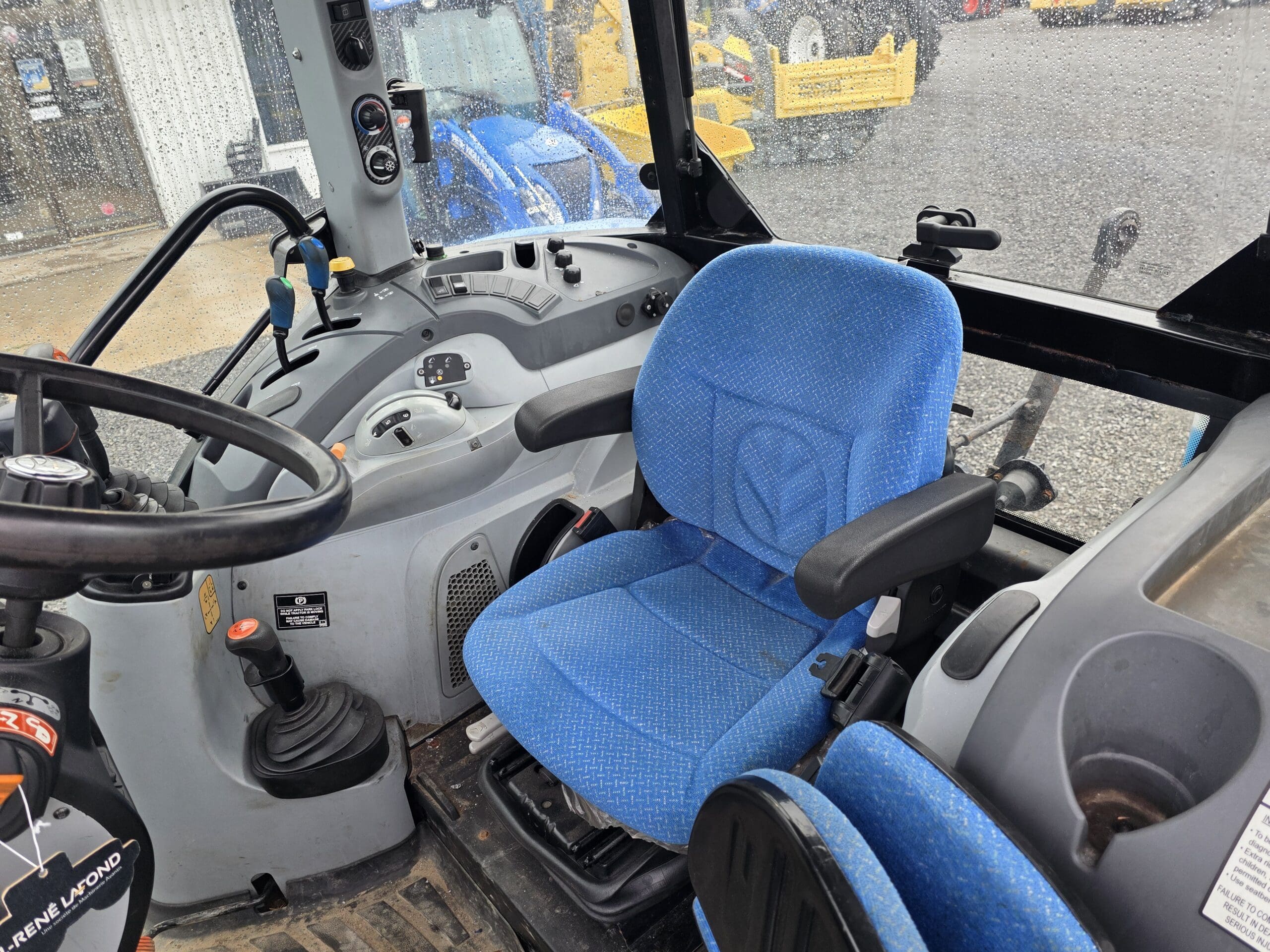 Vue intérieure d'une cabine de tracteur montrant un siège de conducteur en tissu bleu, un volant, divers panneaux de commande et des leviers, avec des gouttes de pluie visibles sur les fenêtres et les machines vues à l'extérieur.