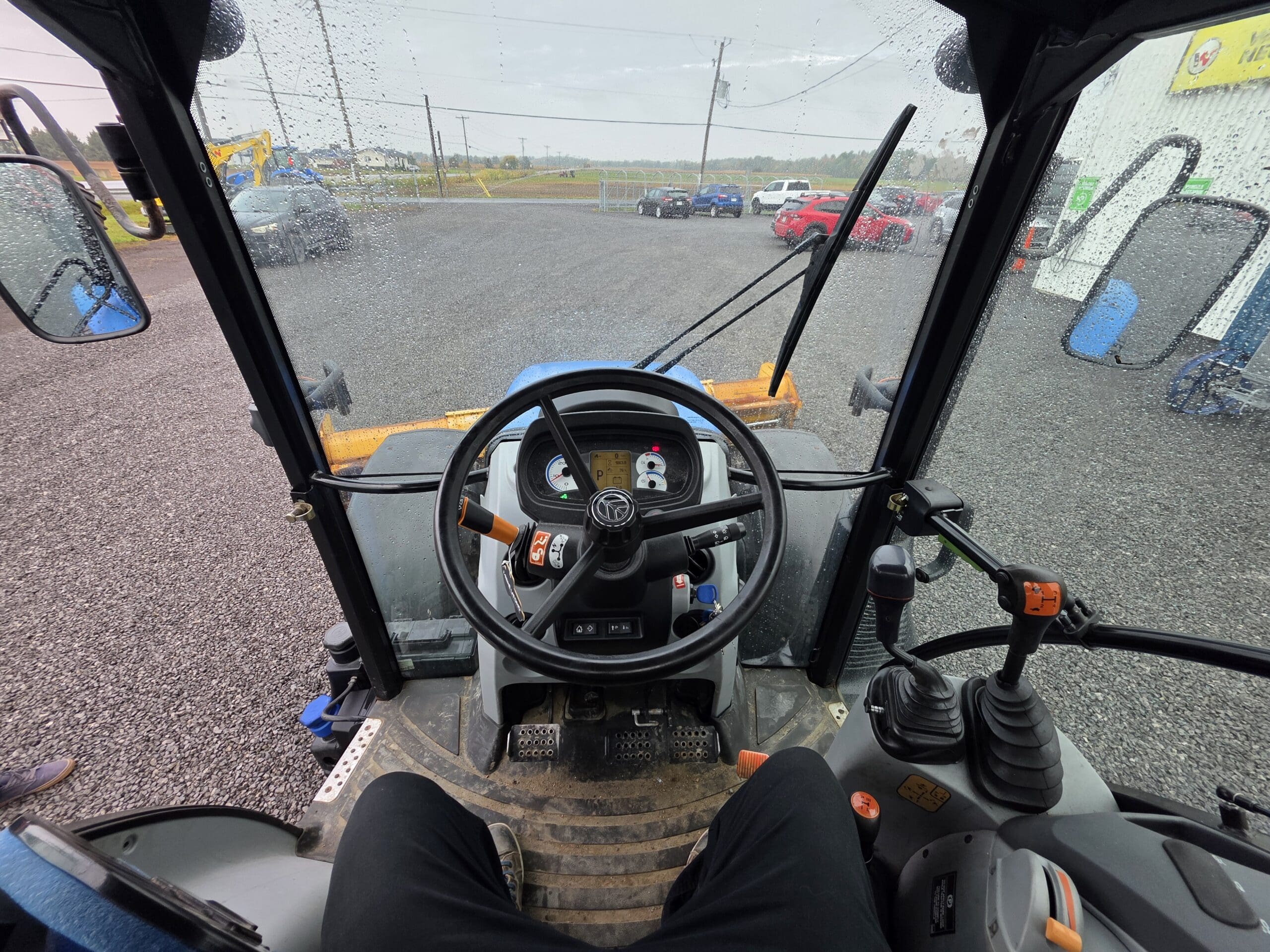 Vue de l'intérieur d'une cabine de tracteur par temps pluvieux, montrant le volant, les commandes du tableau de bord, les pédales, et l'extérieur à travers un pare-brise mouillé sur un terrain en gravier avec des véhicules garés.