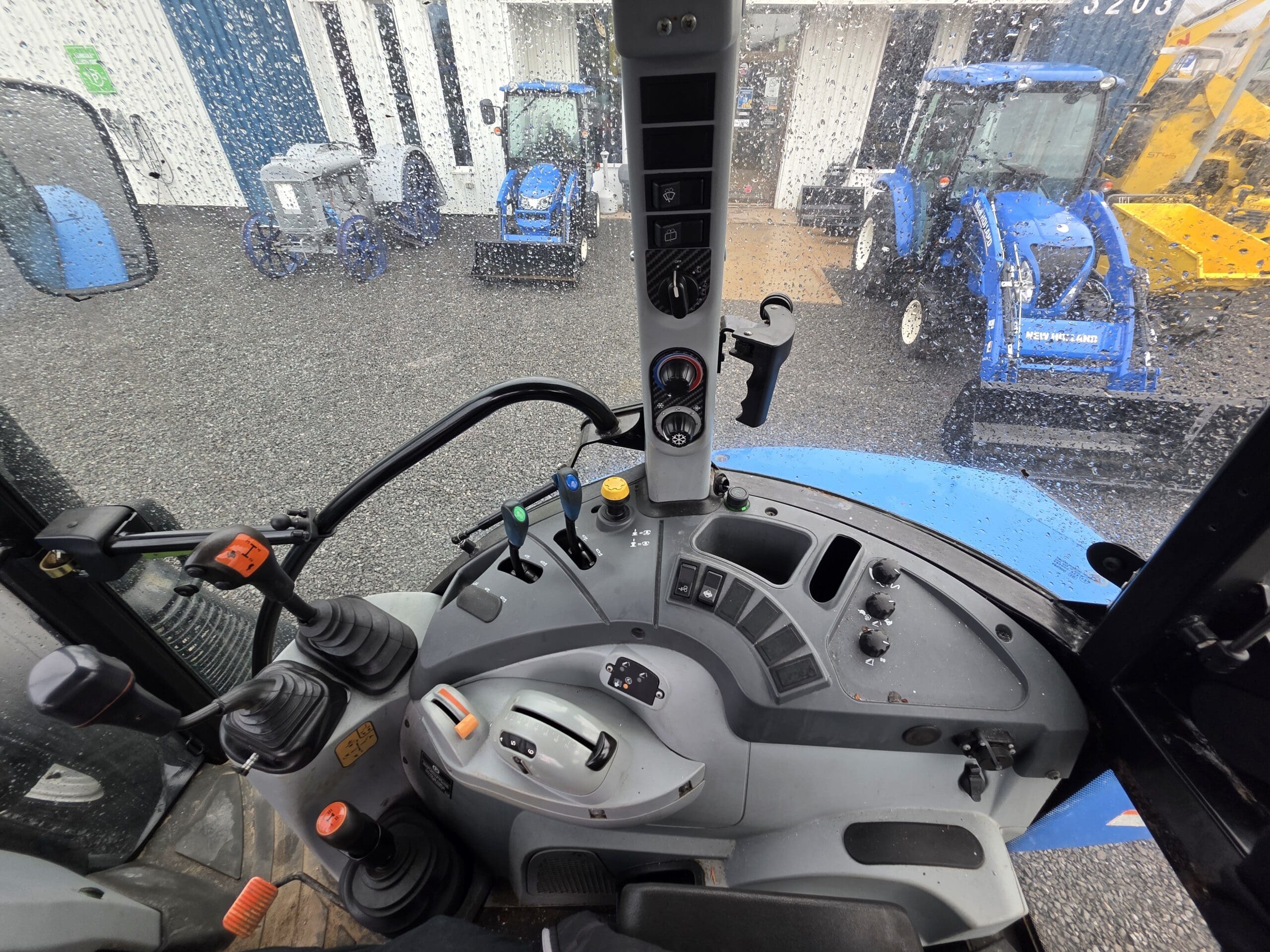 Vue de l'intérieur d'une cabine de tracteur montrant le volant, le tableau de commande, les leviers et les commandes. À l'extérieur, deux tracteurs bleus et d'autres équipements sont visibles à travers un pare-brise tacheté de pluie.