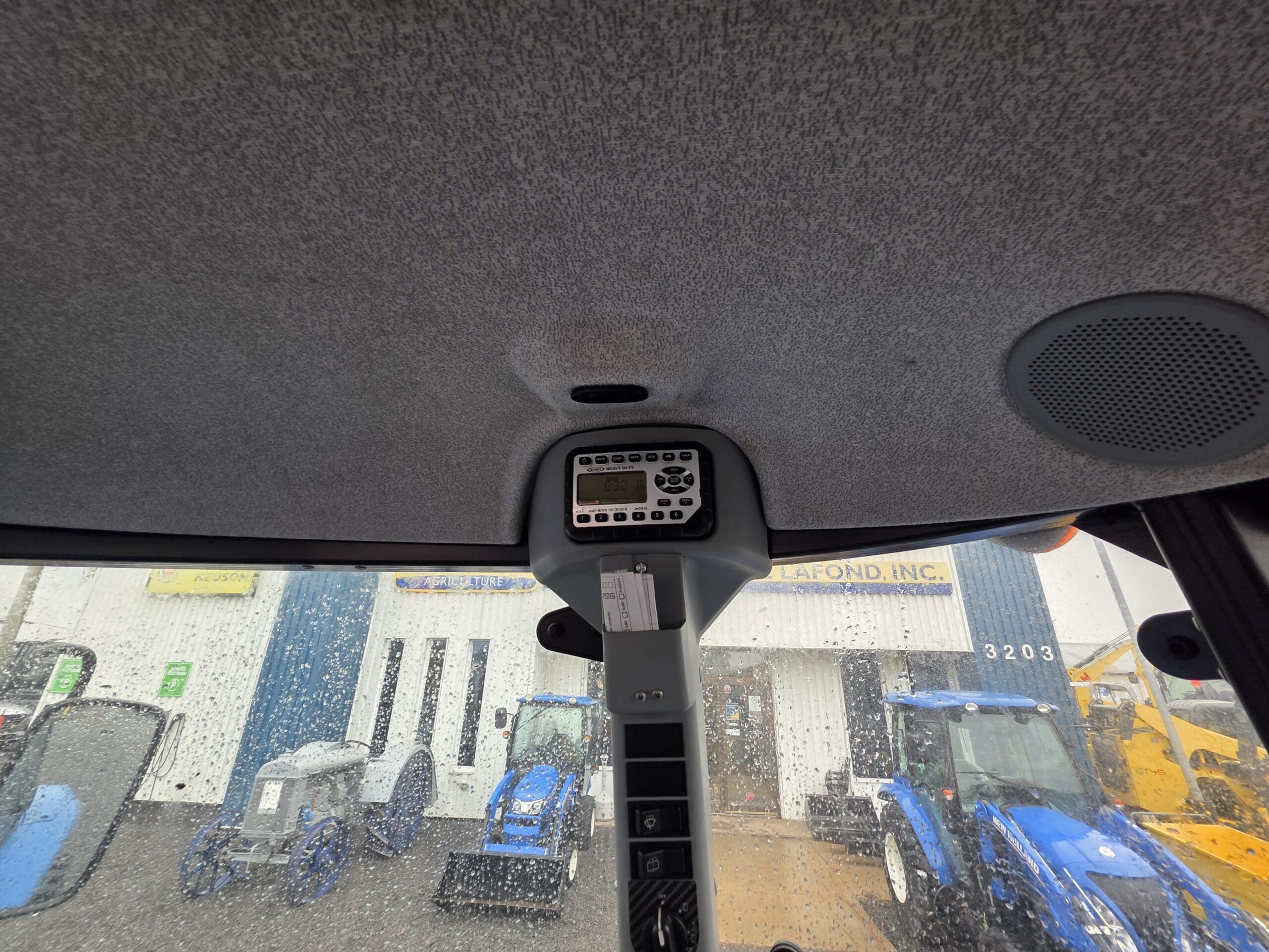 Vue de l'intérieur d'une cabine de tracteur regardant les commandes au plafond, avec une fenêtre pluvieuse montrant des tracteurs bleus et un bâtiment portant l'inscription 