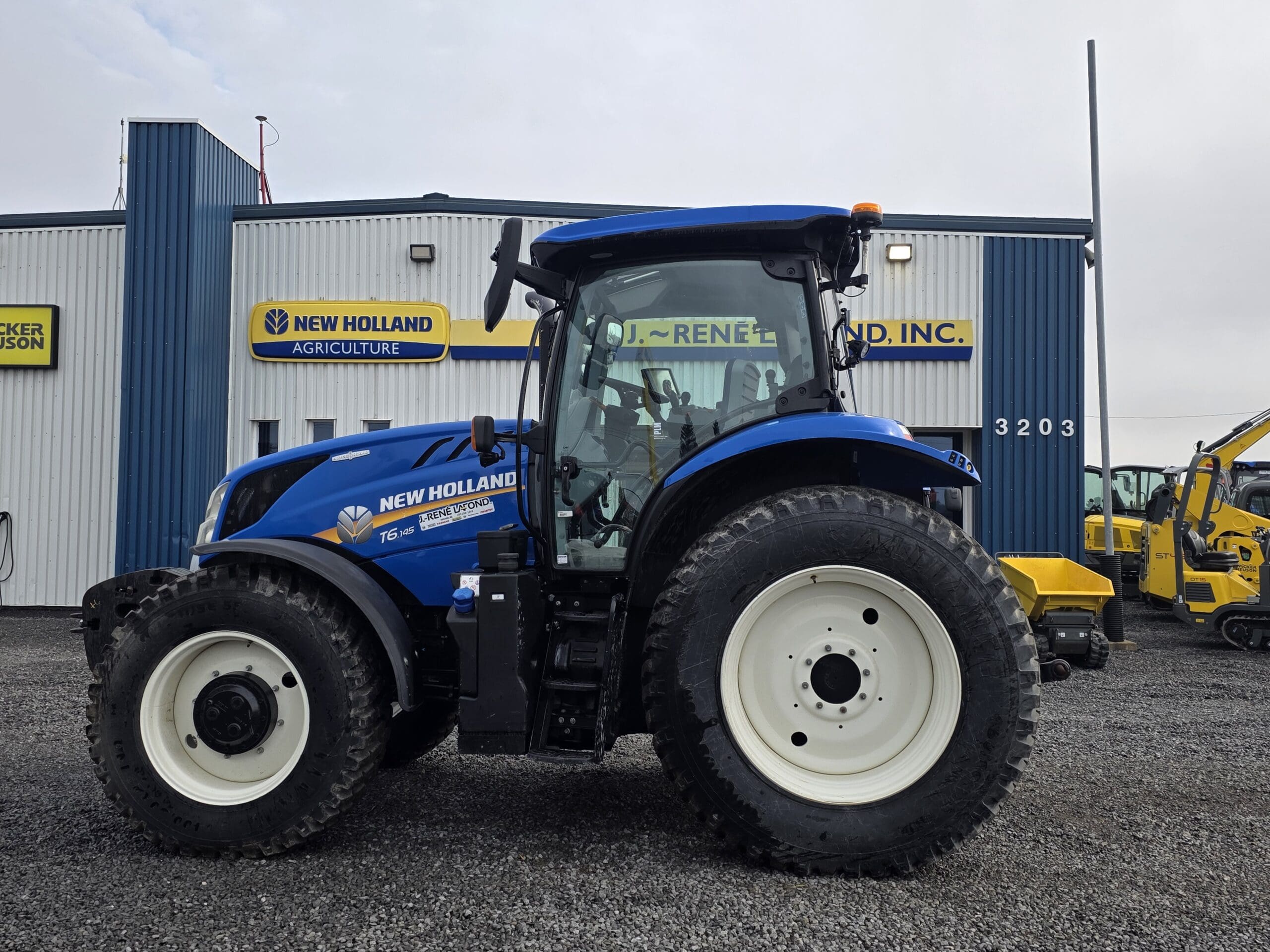 Un tracteur bleu New Holland est garé sur du gravier devant le bâtiment d'un concessionnaire New Holland Agriculture, avec d'autres machines jaunes visibles à l'arrière-plan.