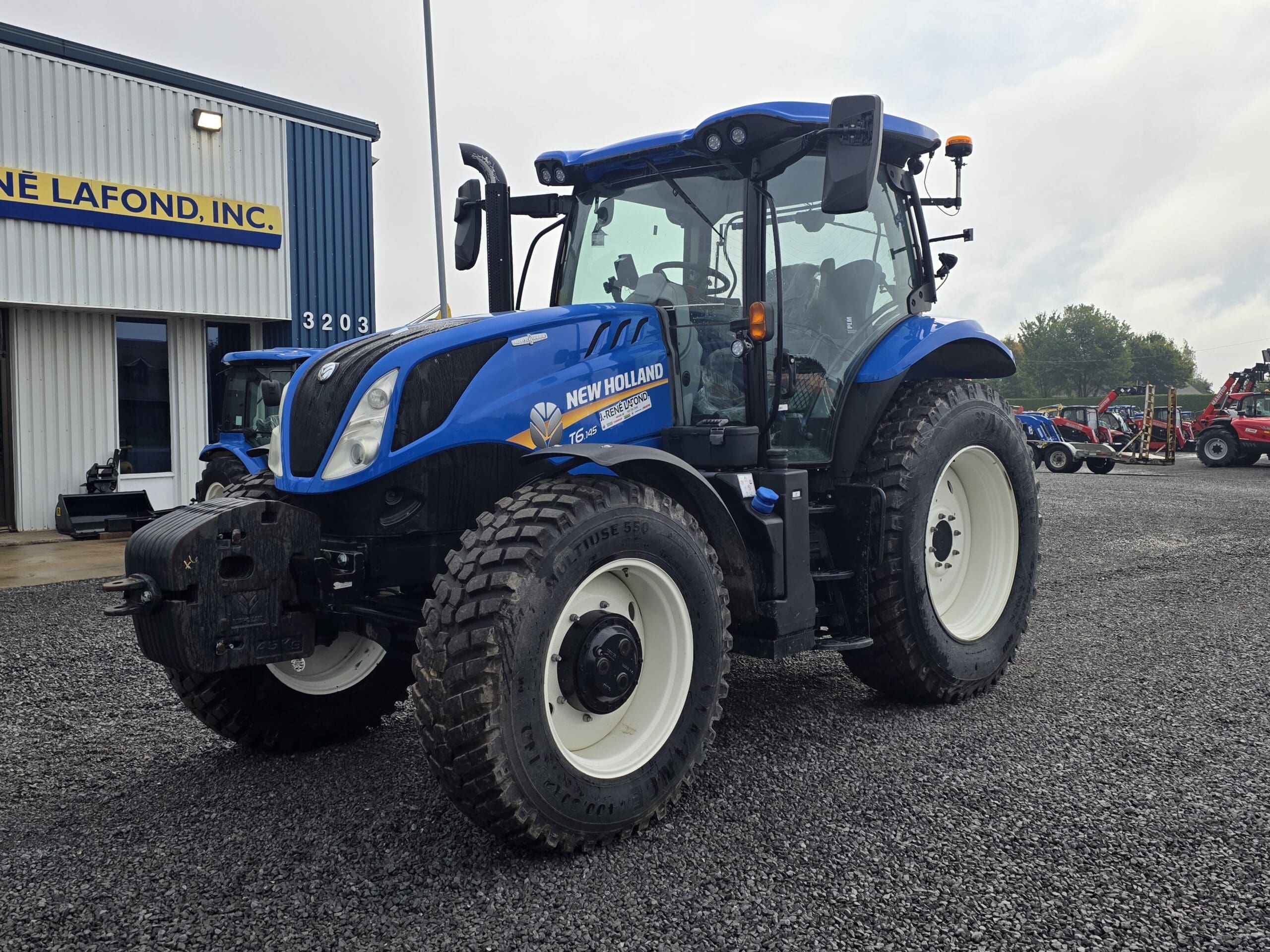 Un tracteur New Holland T6.180 bleu est garé sur un terrain en gravier devant un bâtiment sur lequel on peut lire 