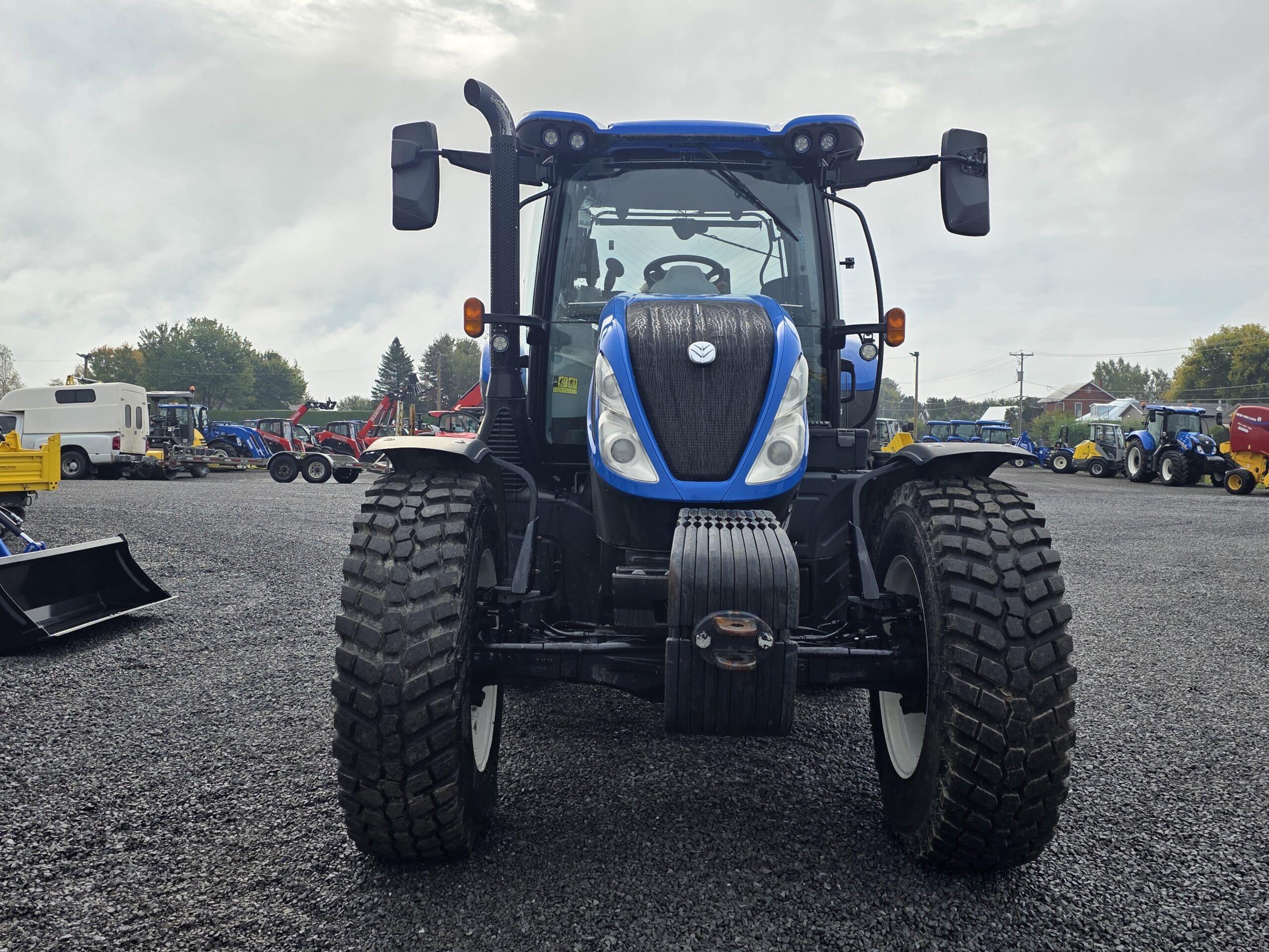 Un tracteur bleu est garé sur du gravier, face à la caméra. D'autres équipements et véhicules agricoles sont visibles à l'arrière-plan sous un ciel nuageux.