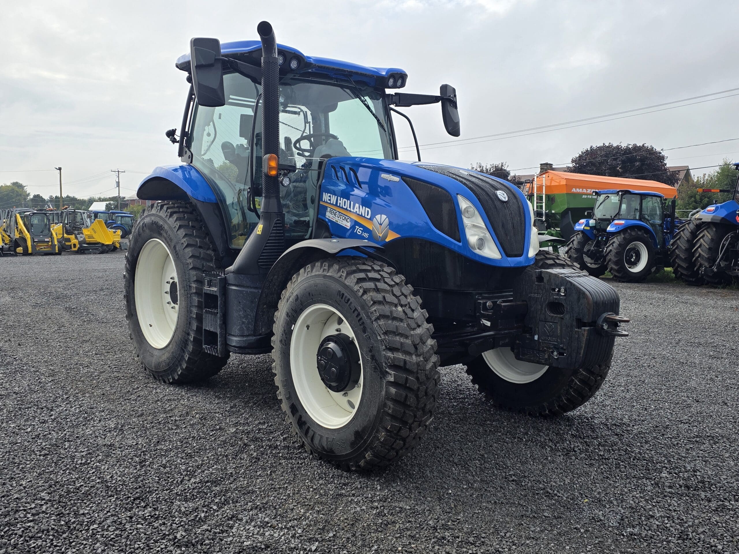 Un tracteur New Holland T6.155 bleu est garé sur un terrain en gravier, entouré d'autres tracteurs et machines, avec un ciel nuageux en arrière-plan.