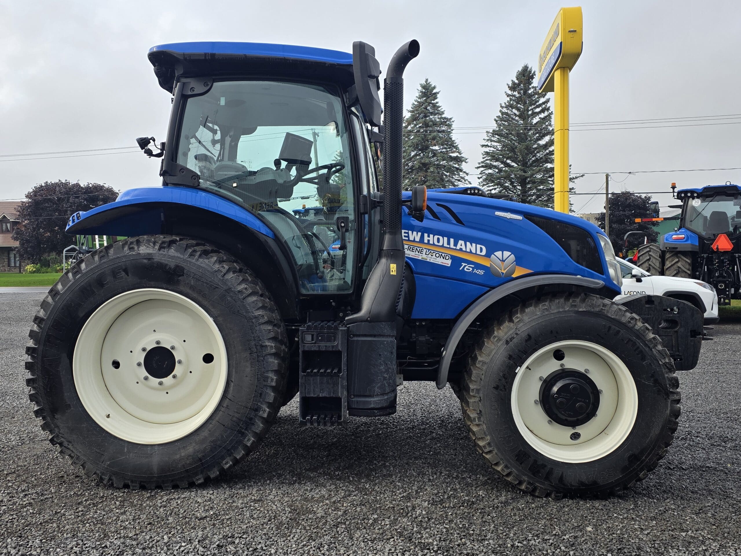 Un tracteur New Holland T5.140 bleu est garé sur un terrain en gravier avec des arbres et un autre tracteur visibles en arrière-plan sous un ciel nuageux.