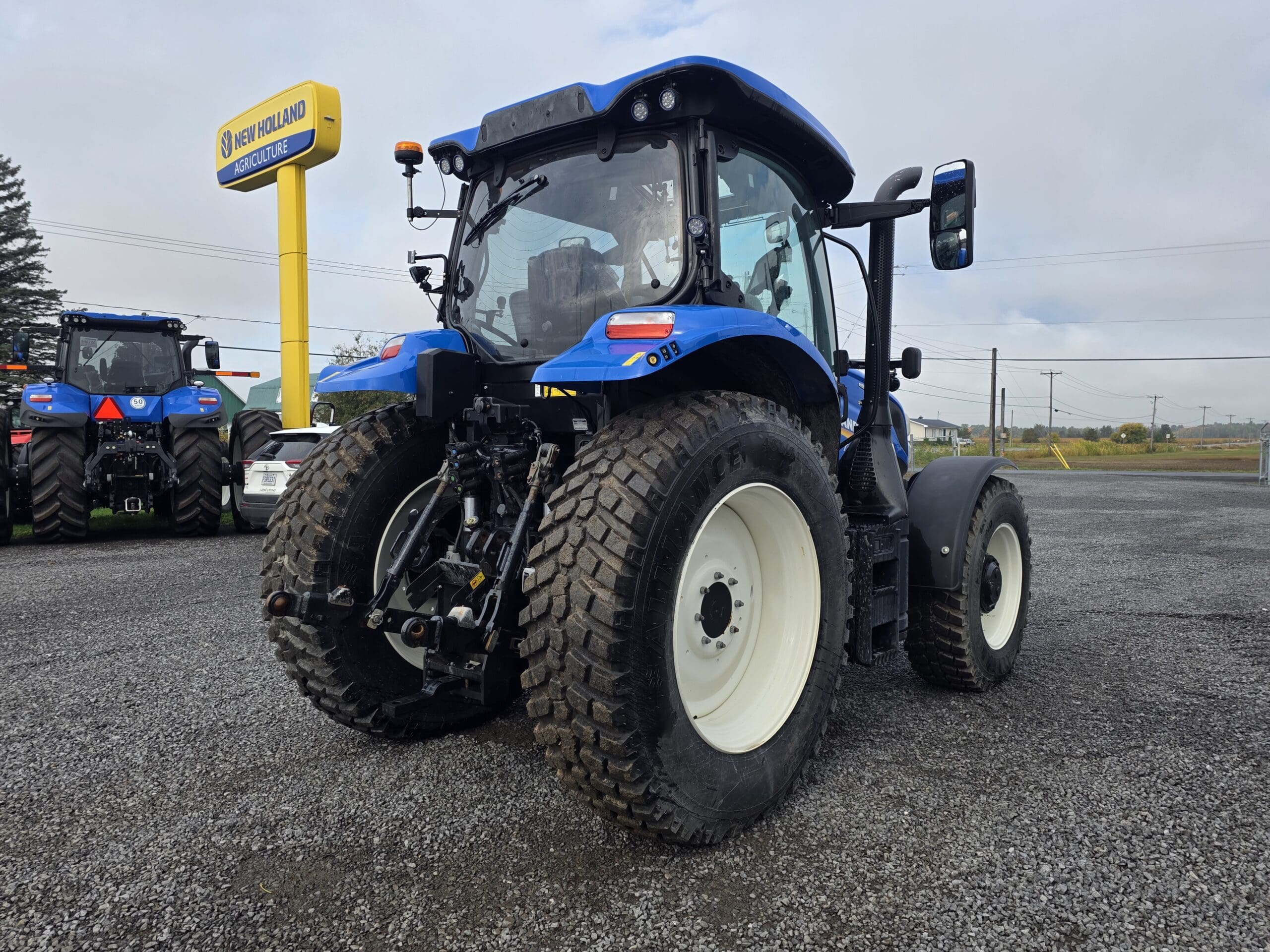 Un tracteur bleu New Holland est garé sur du gravier à l'extérieur d'une concession, avec un autre tracteur et des voitures visibles à l'arrière-plan et un panneau New Holland Agriculture à proximité.