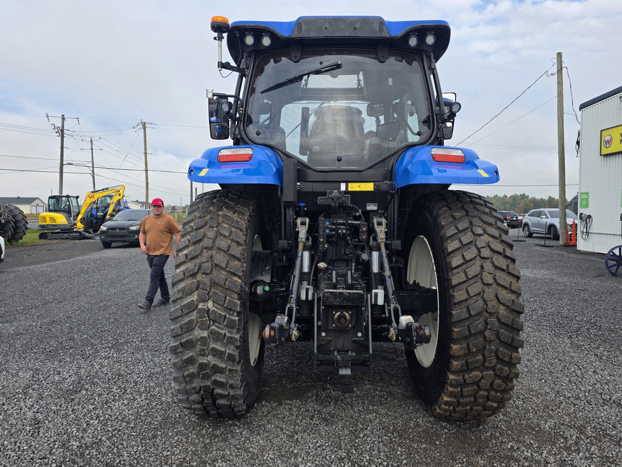Un grand tracteur bleu vu de l'arrière, garé sur du gravier à côté de bâtiments industriels. Une personne vêtue d'une chemise brune et d'une casquette rouge marche à proximité, et l'on aperçoit d'autres machines à l'arrière-plan.