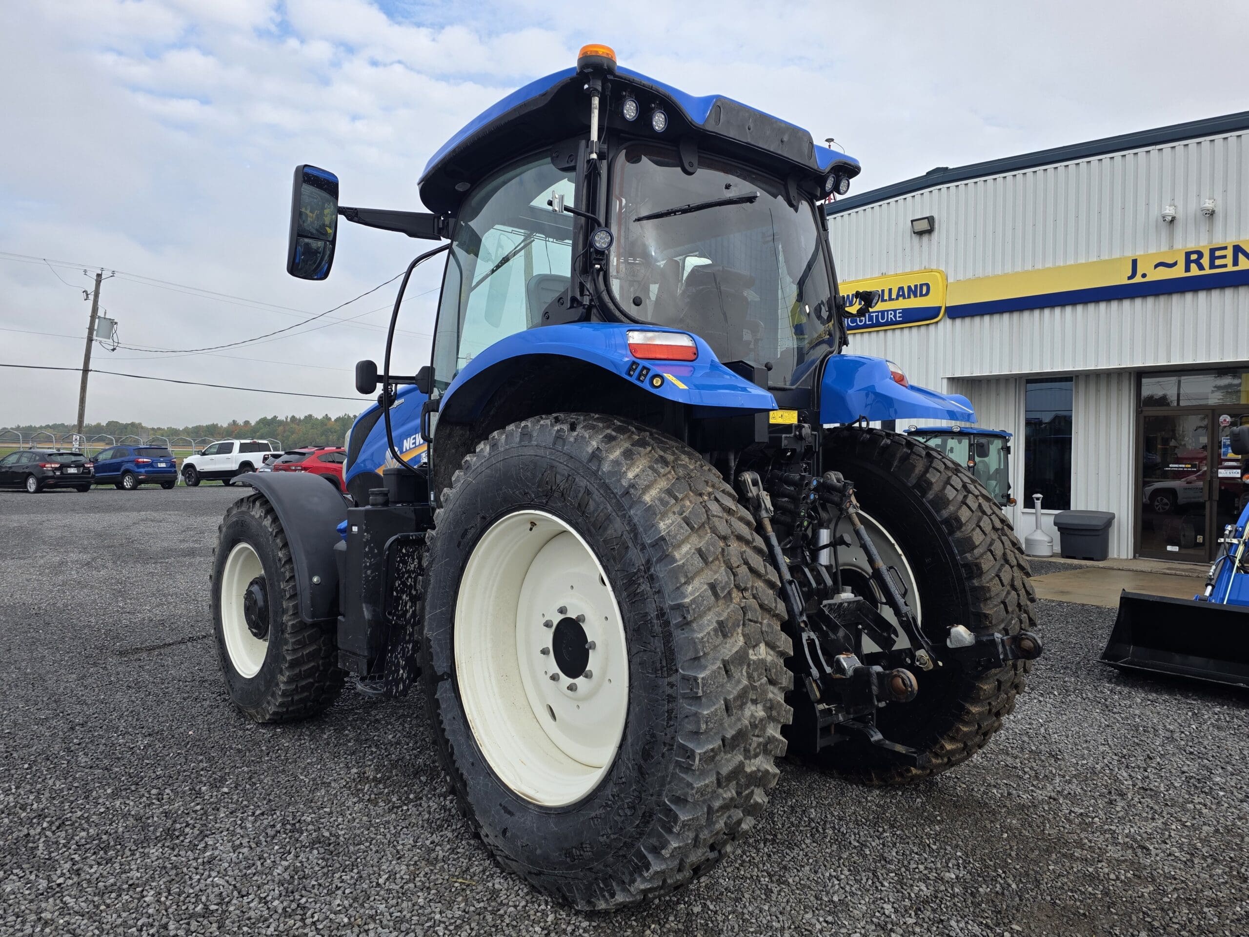 Un grand tracteur bleu aux pneus robustes est garé sur du gravier à l'extérieur d'un bâtiment portant une enseigne jaune et bleue indiquant 