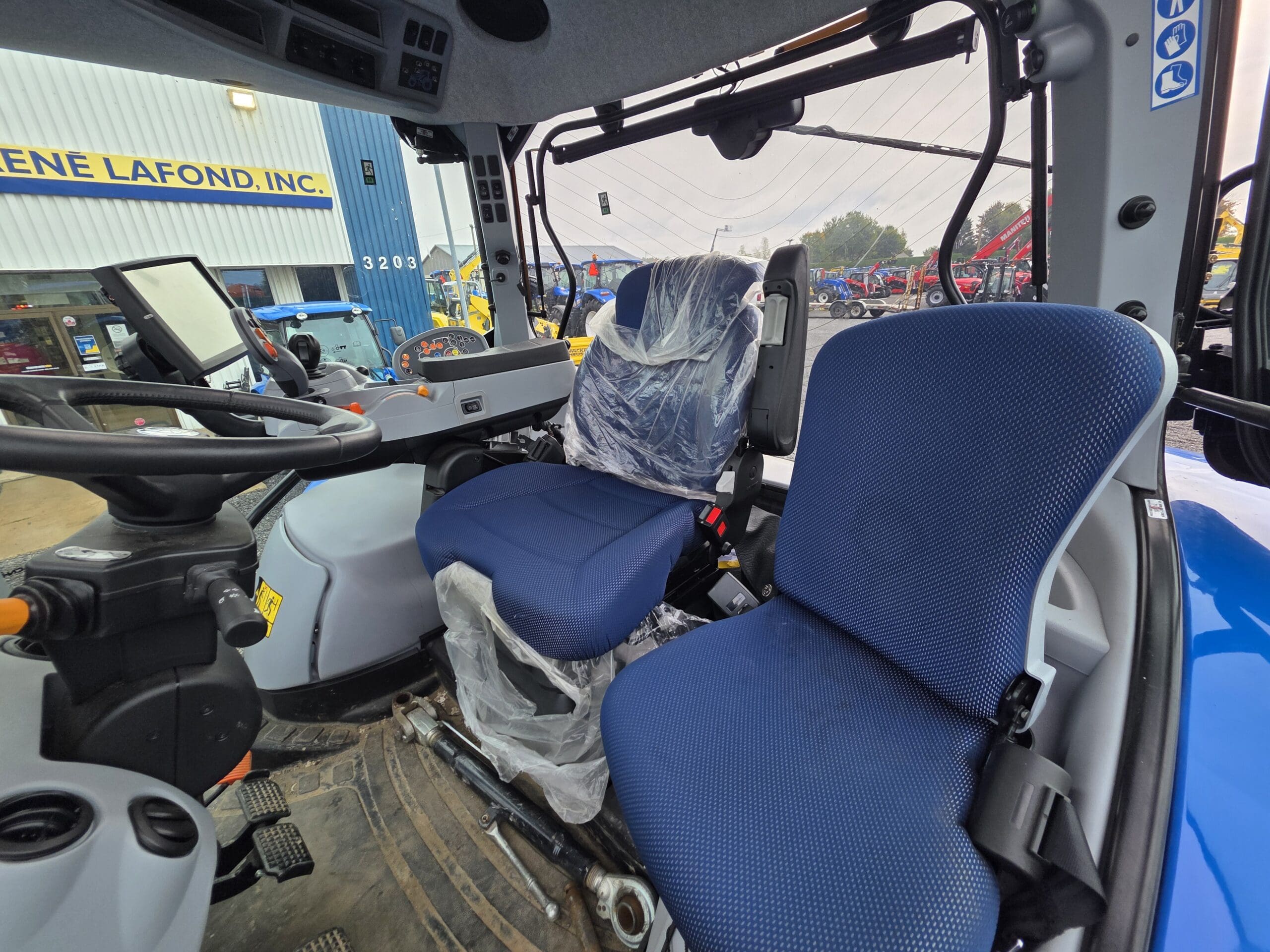 Vue intérieure d'une cabine de tracteur moderne bleue avec deux sièges bleus, dont l'un est recouvert de plastique, un volant, des commandes et l'extérieur d'un concessionnaire visible à travers les fenêtres.