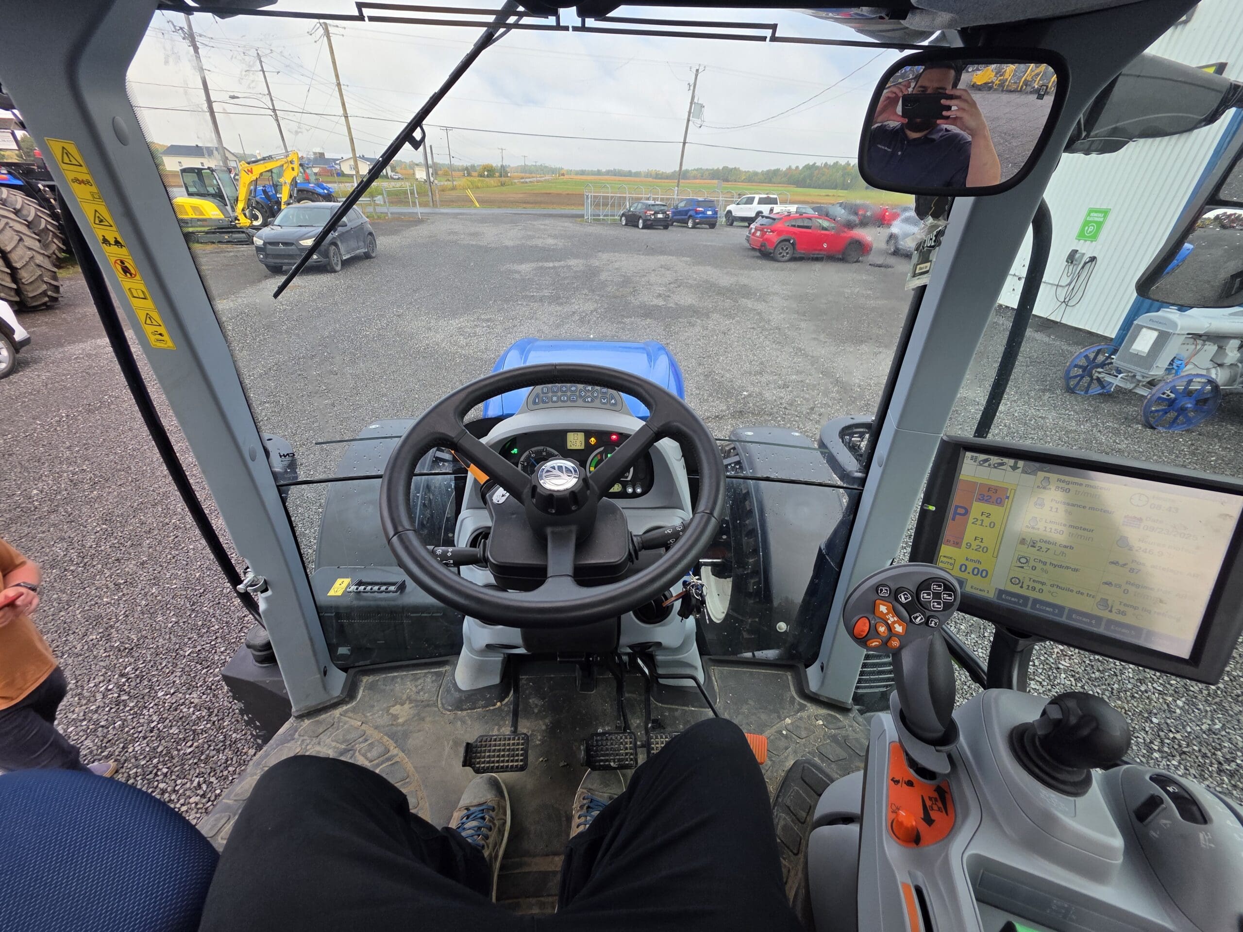 Vue de l'intérieur d'une cabine de tracteur montrant le volant, les pédales, l'écran d'affichage numérique, divers boutons de commande et un terrain extérieur avec des voitures, des machines et des champs visibles à travers le pare-brise.
