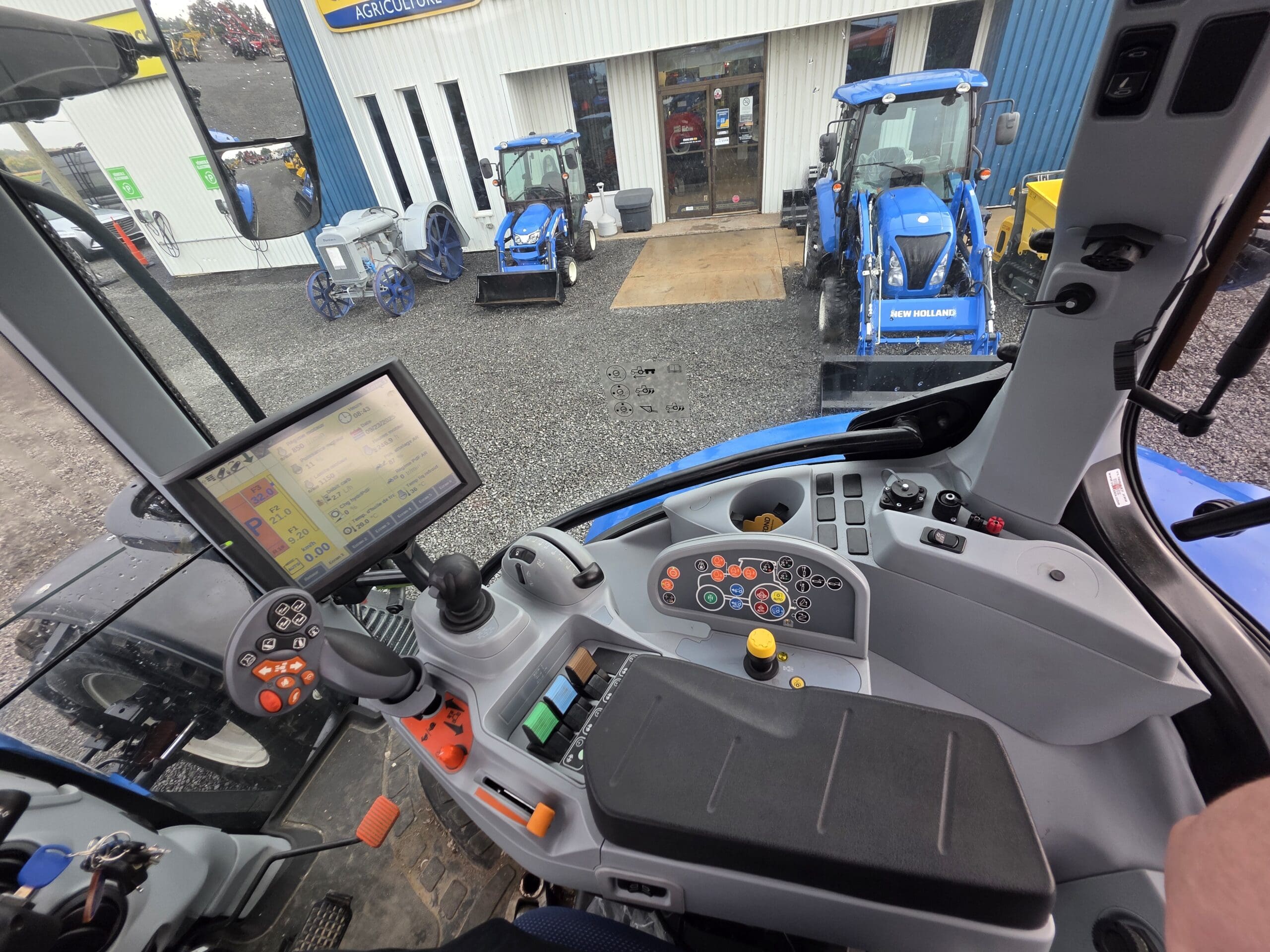 Vue de l'intérieur d'une cabine de tracteur montrant les commandes, l'écran et le volant, avec plusieurs tracteurs bleus garés à l'extérieur d'un bâtiment visible à travers le pare-brise.