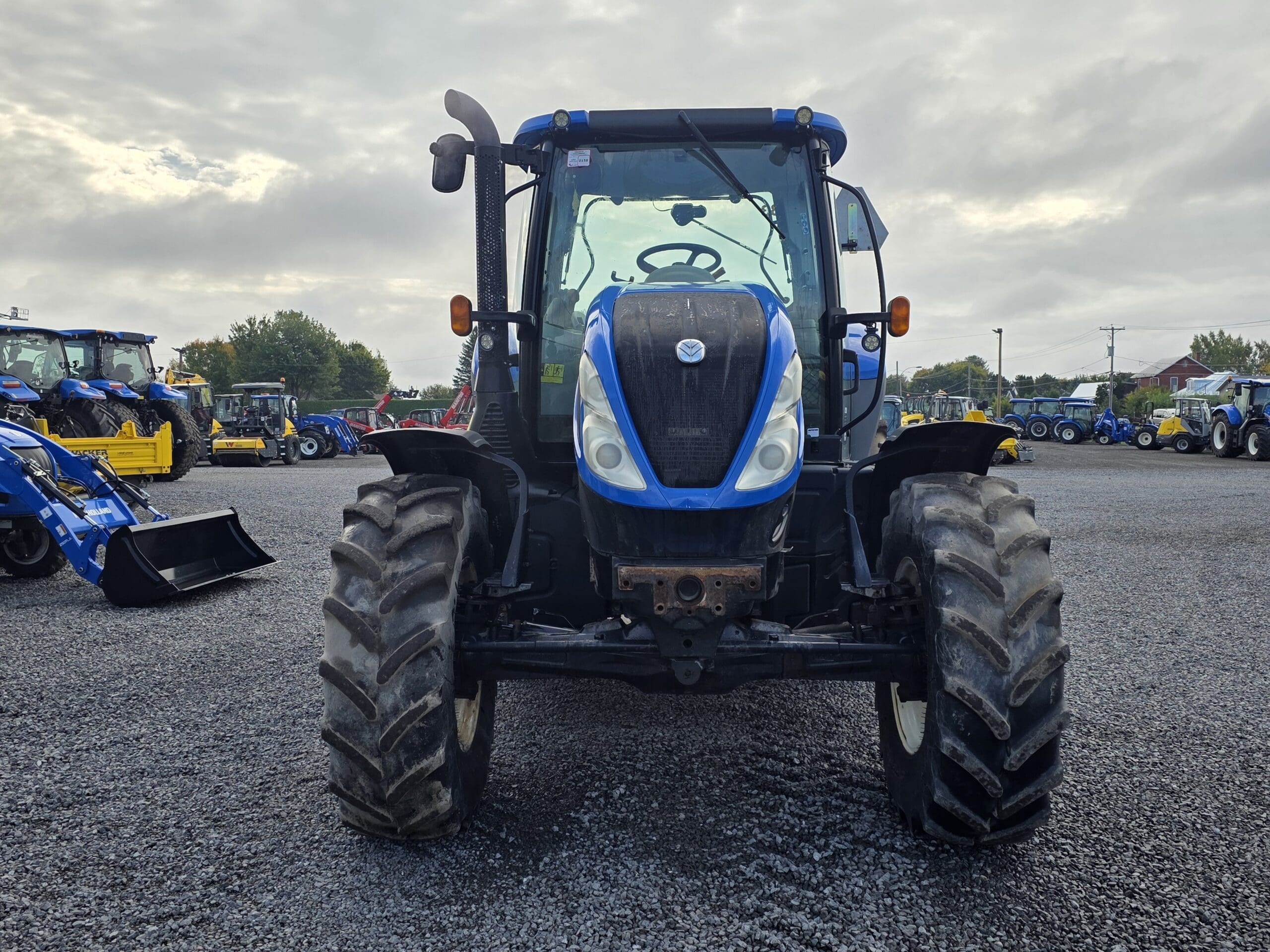 Un tracteur bleu et noir est garé sur un terrain en gravier, vu de face, avec d'autres tracteurs et équipements visibles en arrière-plan sous un ciel nuageux.