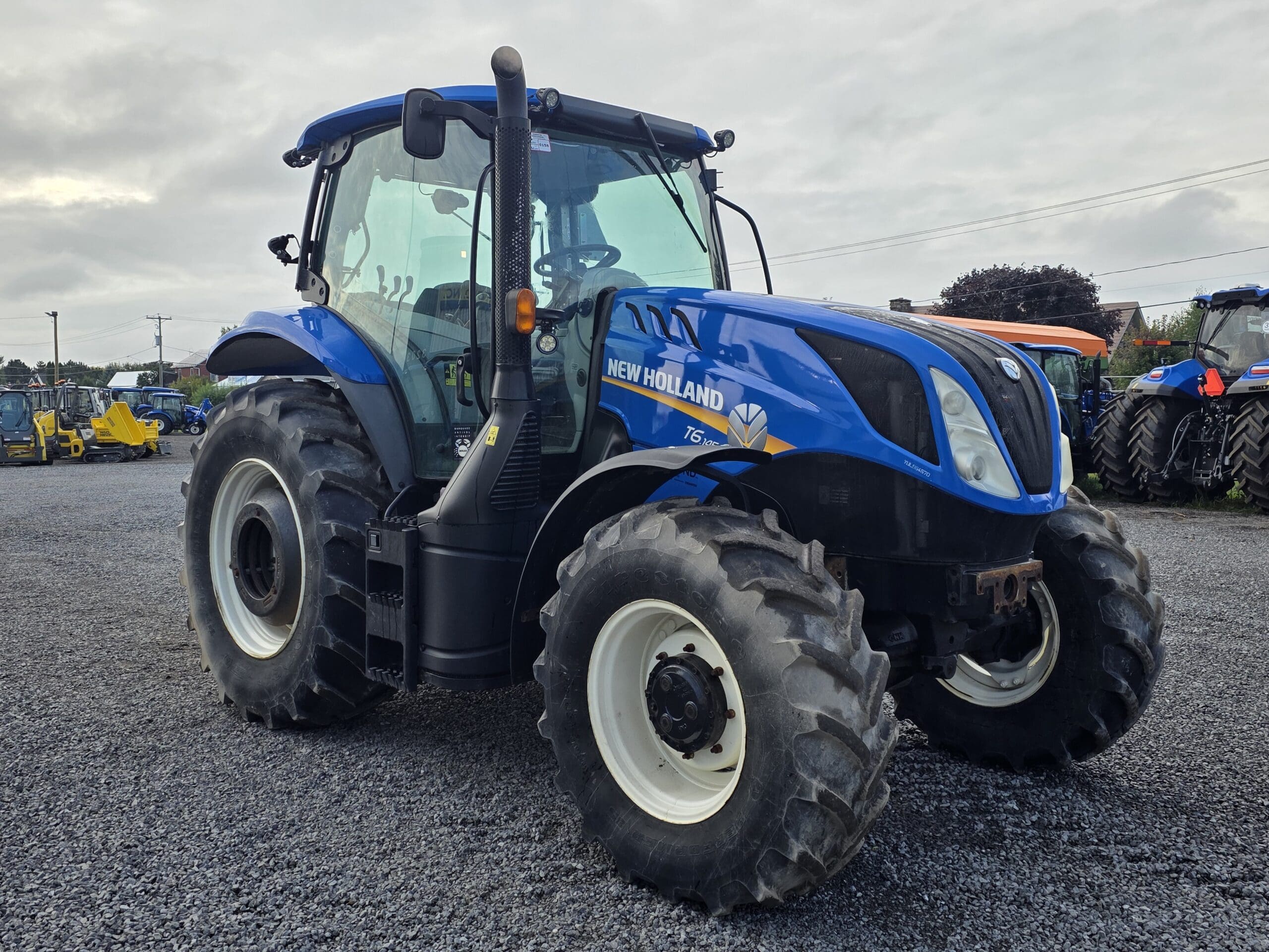 Un tracteur New Holland bleu à gros pneus est garé sur du gravier, avec un ciel nuageux et d'autres équipements agricoles visibles à l'arrière-plan.