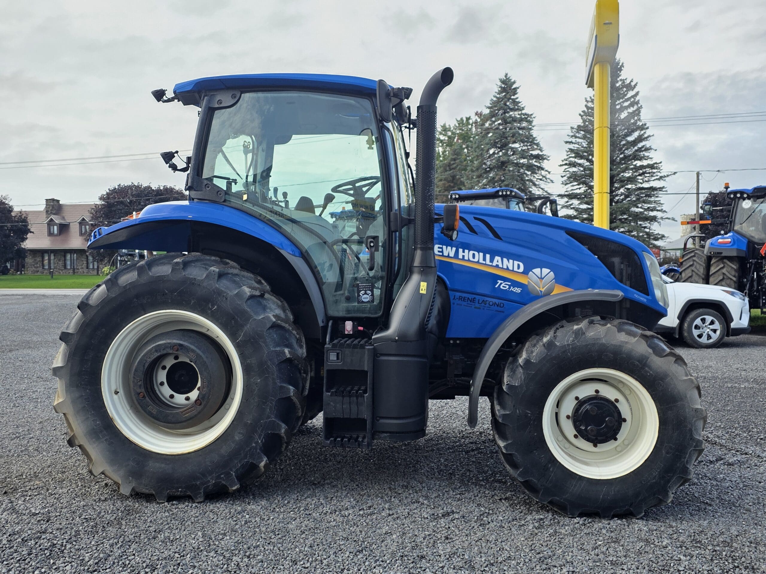 Un tracteur New Holland T5.95 bleu avec de gros pneus est garé sur un terrain en gravier, avec des arbres, un bâtiment et d'autres véhicules visibles à l'arrière-plan.