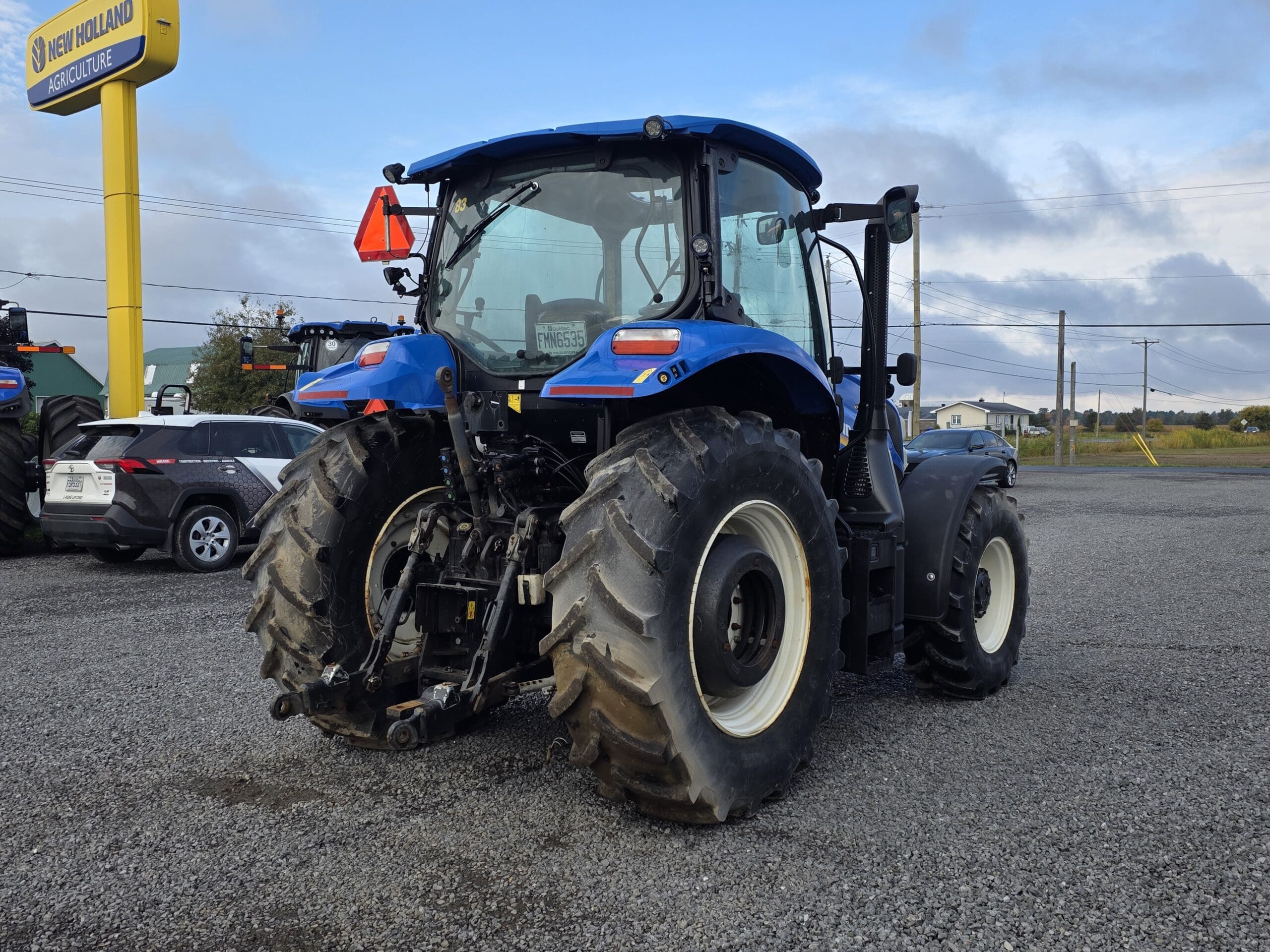 Un tracteur bleu New Holland est garé sur un terrain en gravier près d'un panneau New Holland Agriculture. Plusieurs véhicules sont visibles à l'arrière-plan sous un ciel nuageux.