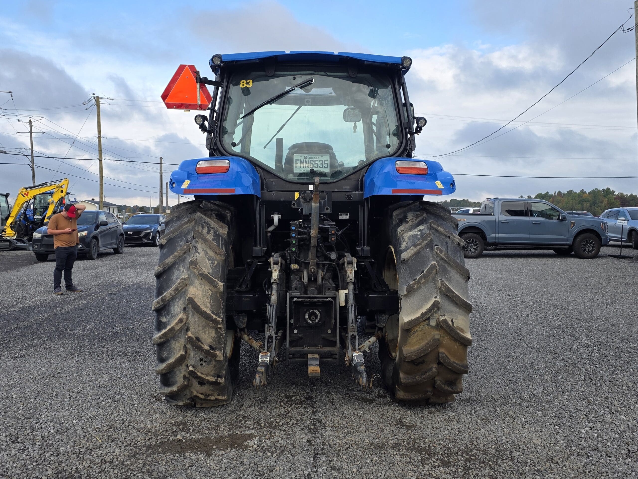 Un tracteur bleu vu de l'arrière, garé sur du gravier, avec plusieurs voitures et une personne se tenant à proximité dans un terrain sous un ciel nuageux.