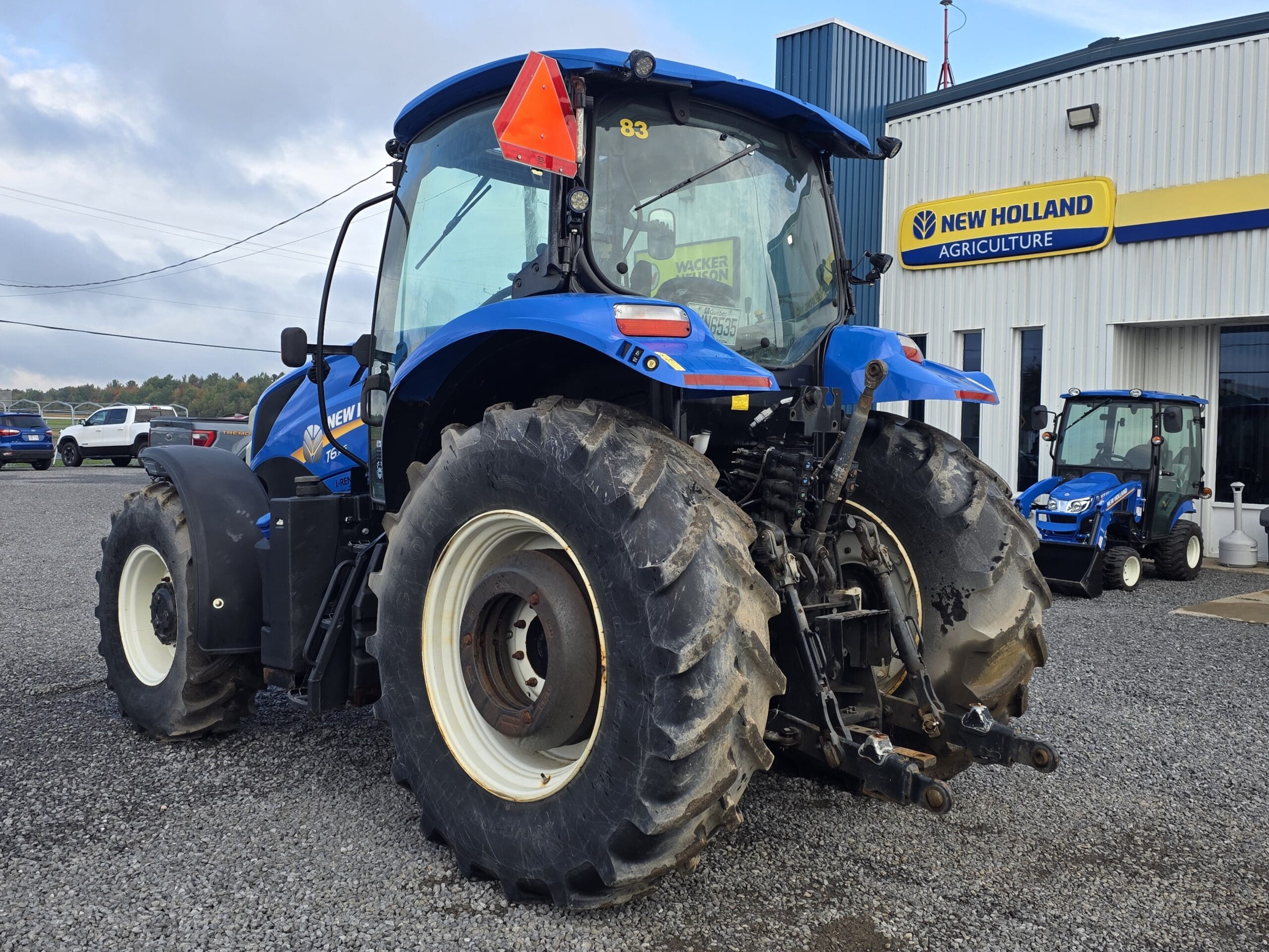 Un grand tracteur bleu New Holland est garé sur du gravier près du bâtiment d'un concessionnaire New Holland Agriculture. Un autre tracteur bleu plus petit est visible près de l'entrée du bâtiment.