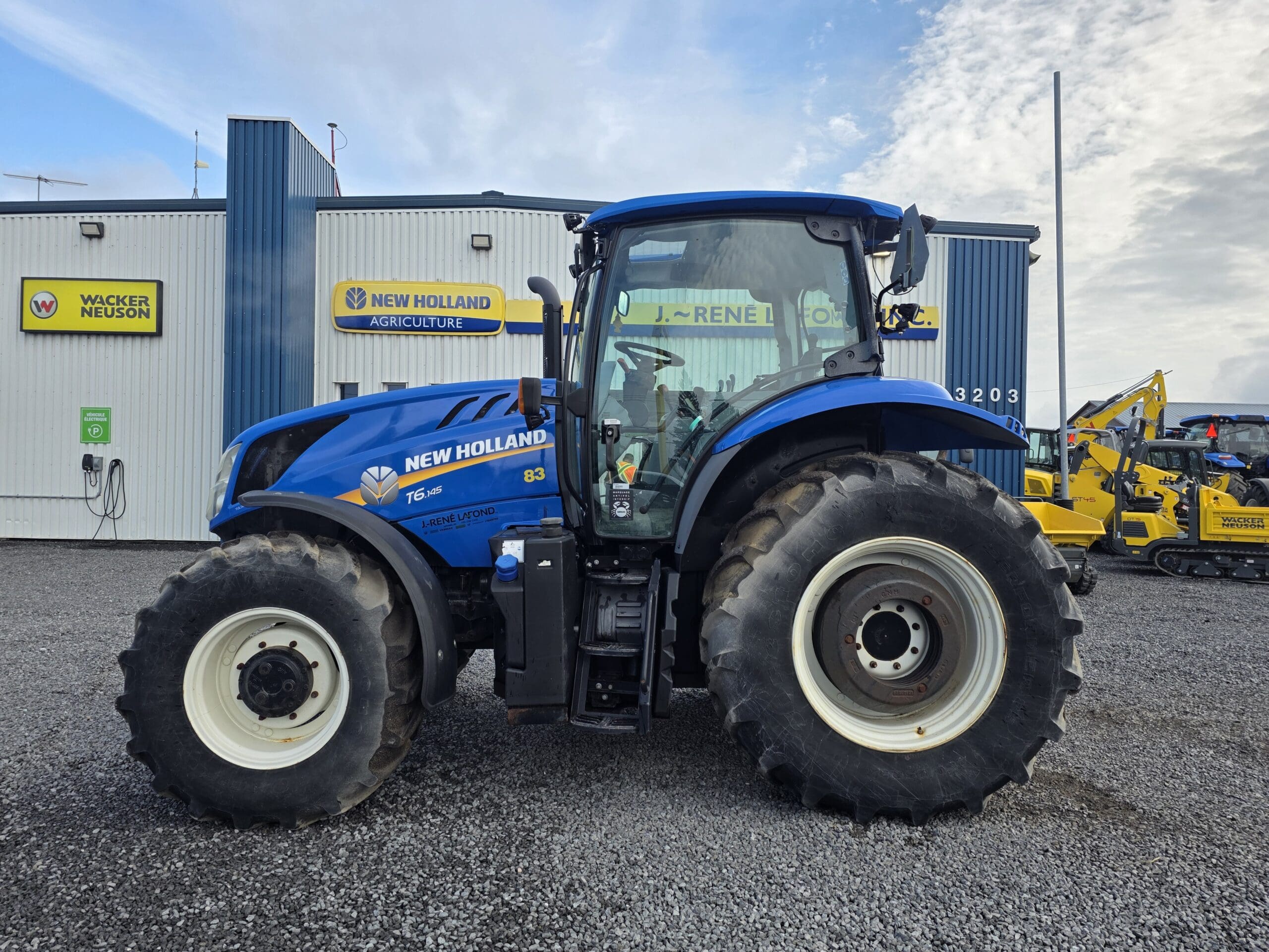 Un tracteur bleu New Holland T6.160 est garé sur du gravier devant un bâtiment avec des panneaux New Holland Agriculture et d'autres équipements de construction visibles à l'arrière-plan.