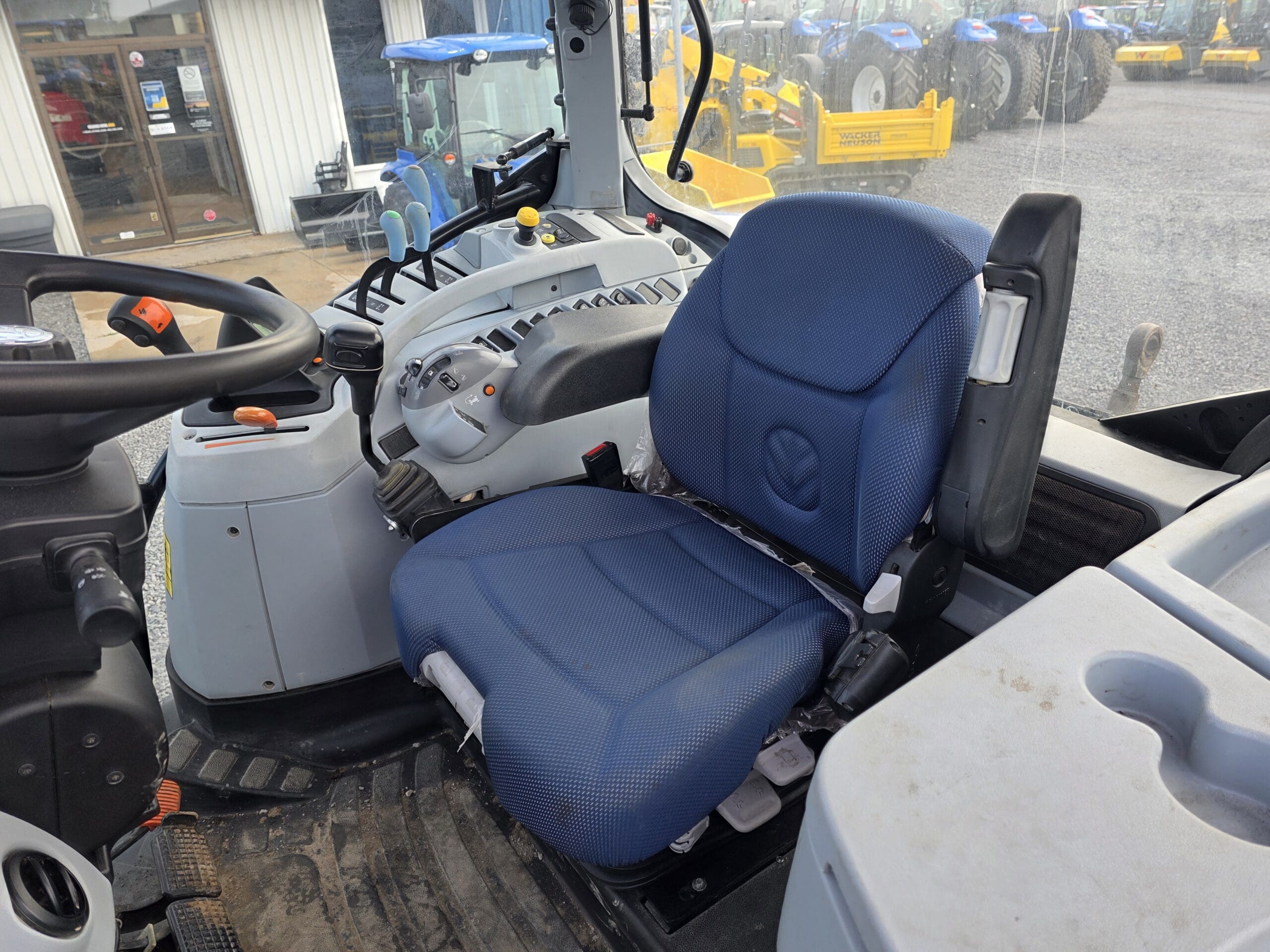 Siège du conducteur et tableau de commande à l'intérieur d'une cabine de tracteur moderne, comprenant un siège à coussin bleu, divers leviers et boutons, ainsi qu'une partie du volant, les tracteurs étant visibles par la fenêtre à l'extérieur.