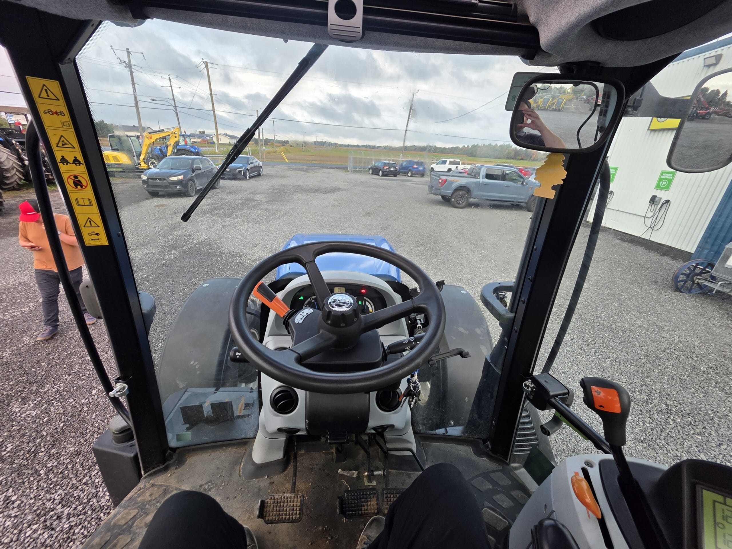 Vue de l'intérieur d'une cabine de tracteur montrant le volant, le tableau de bord et les leviers de commande. À l'extérieur, plusieurs voitures garées, du matériel et quelques personnes sont visibles sur un terrain en gravier sous un ciel nuageux.